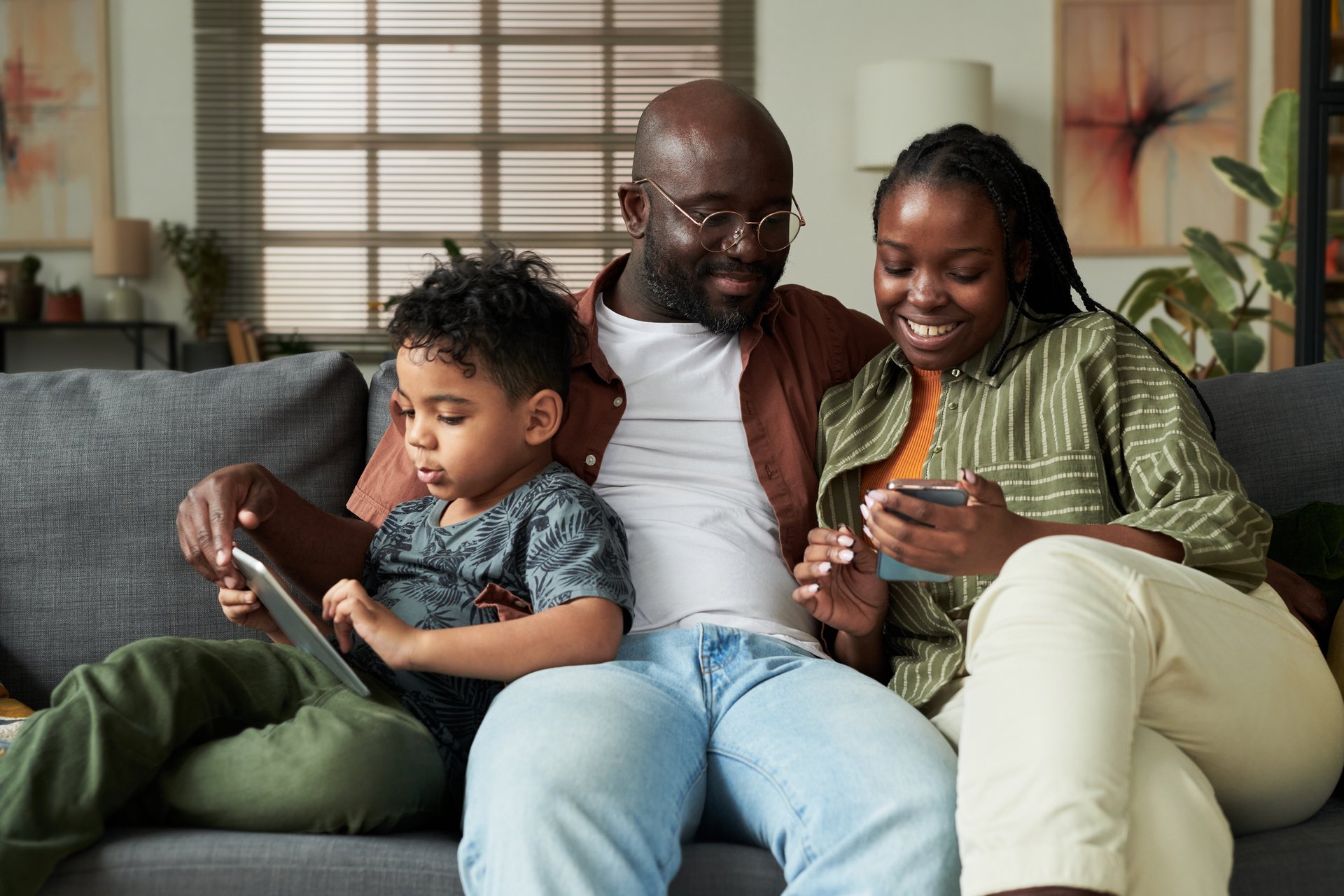 Young African American couple looking at screen of smartphone while sitting next to their son with touchpad watching online video