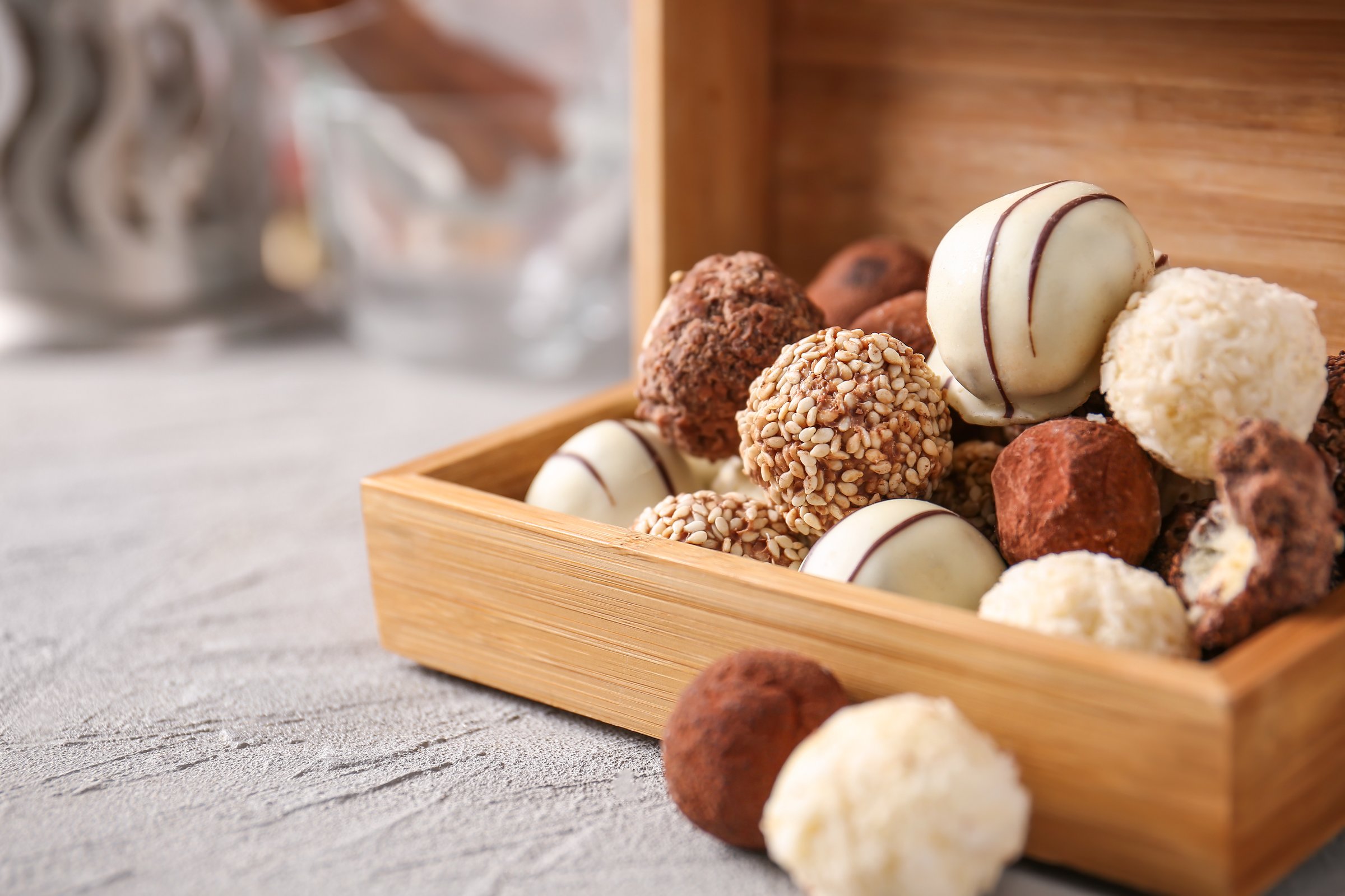 Appetizing still life photograph of gourmet assorted chocolate truffles artfully arranged in a rustic wooden crate, highlighting various textures and coatings?from smooth white chocolate drizzle to crunchy sesame seeds and shredded coconut. The candies sit on a neutral gray concrete background, lending an elegant, rustic mood. Perfect for Valentine's Day, holiday campaigns, or confectionery product advertising.