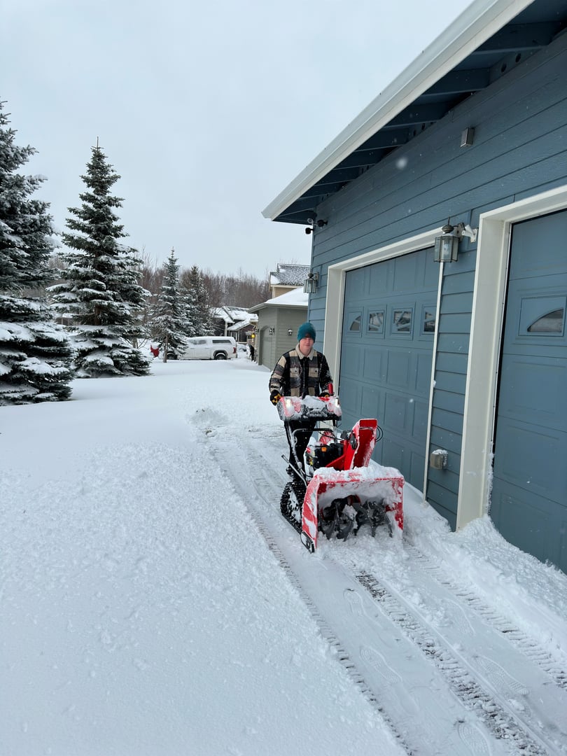 Residential snow removal service in Anchorage, Alaska, featuring a worker operating a snowblower in a snowy driveway, surrounded by evergreen trees and a blue garage.