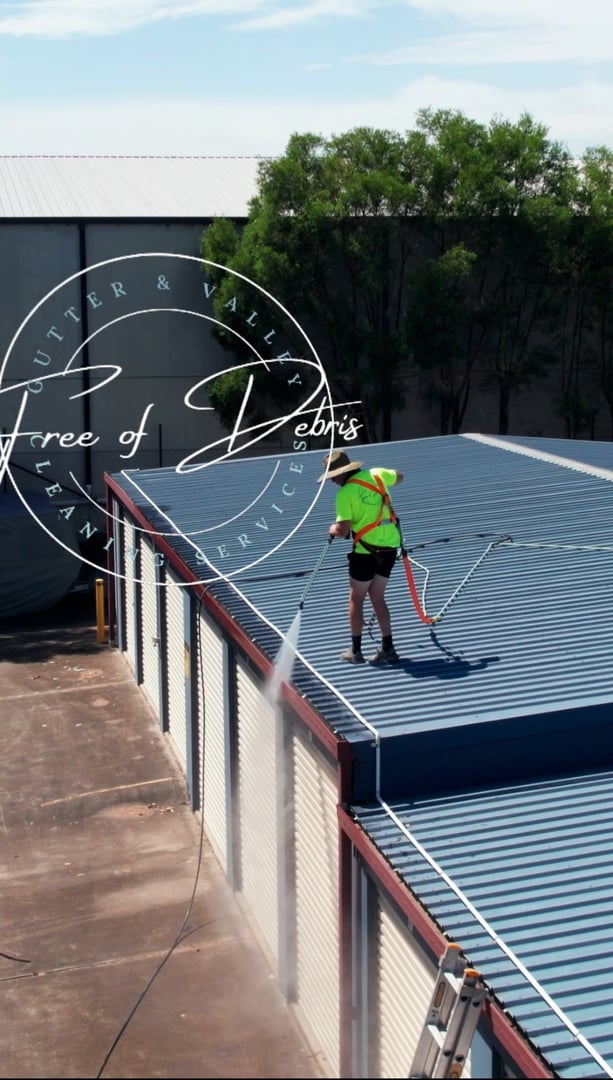 Person in safety gear pressure washing a blue metal roof, surrounded by industrial buildings and trees.