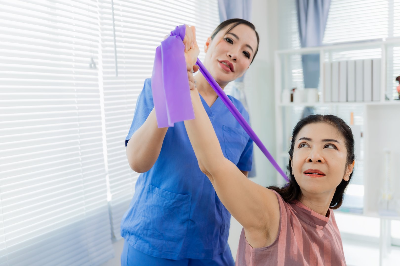 An Asian nurse assists a patient with exercise using resistance bands during physical therapy, with the nurse providing assistance in the patient's room.