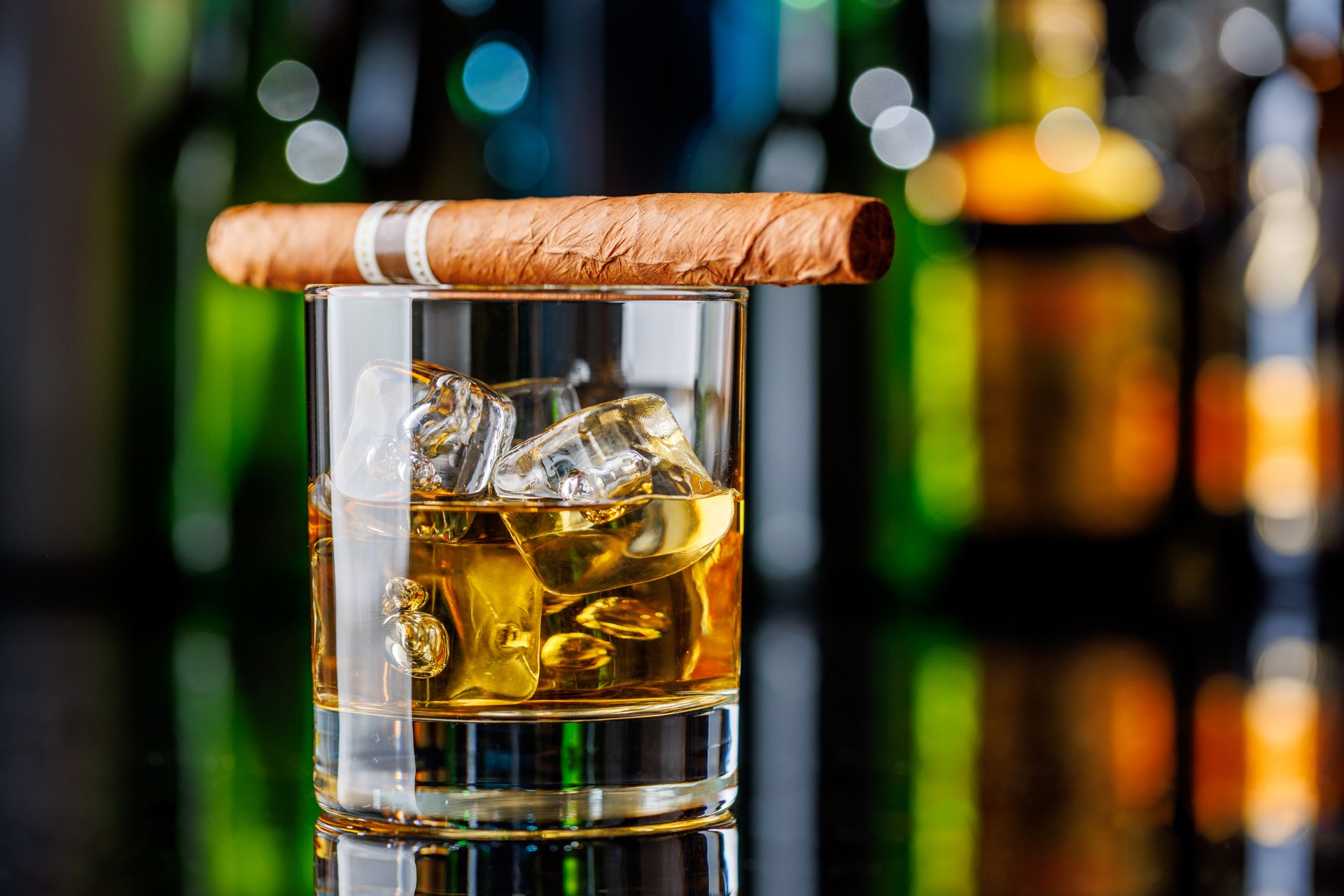 Whiskey on the rocks beside a lit cigar on a bar table. The backdrop features colorful bokeh from an array of illuminated bottles, adding depth and ambiance