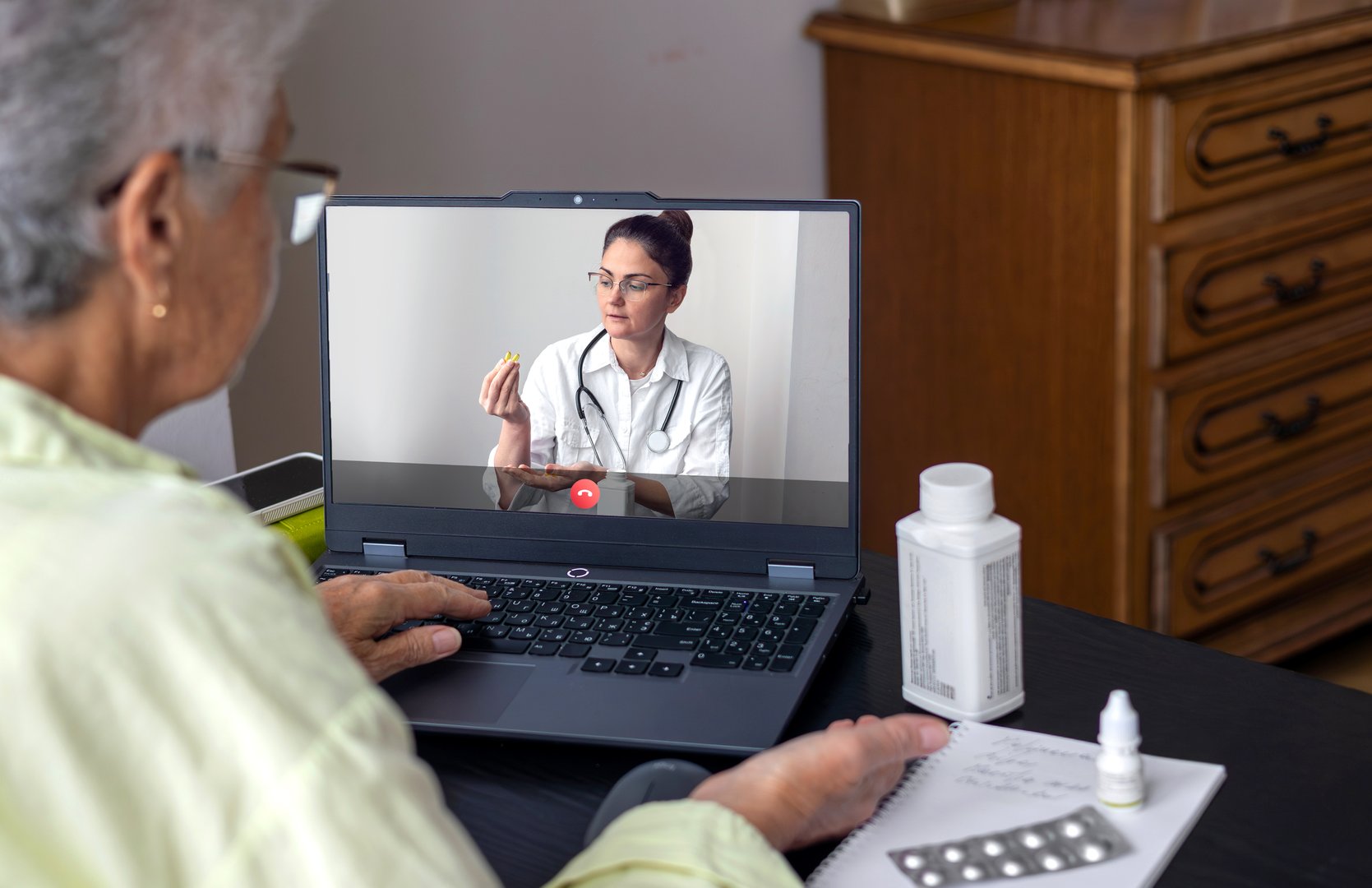 An elderly woman sits at a table in front of her laptop, participating in a video consultation with a health professional (doctor, dietitian or nutritionist). She holds a handful of transparent yellow supplement capsules (such as Omega-3 or Vitamin D) emphasized in her hand while attentively listening on screen. The scene highlights modern telemedicine, active senior wellness, prevention of age-related conditions and digital healthcare engagement.