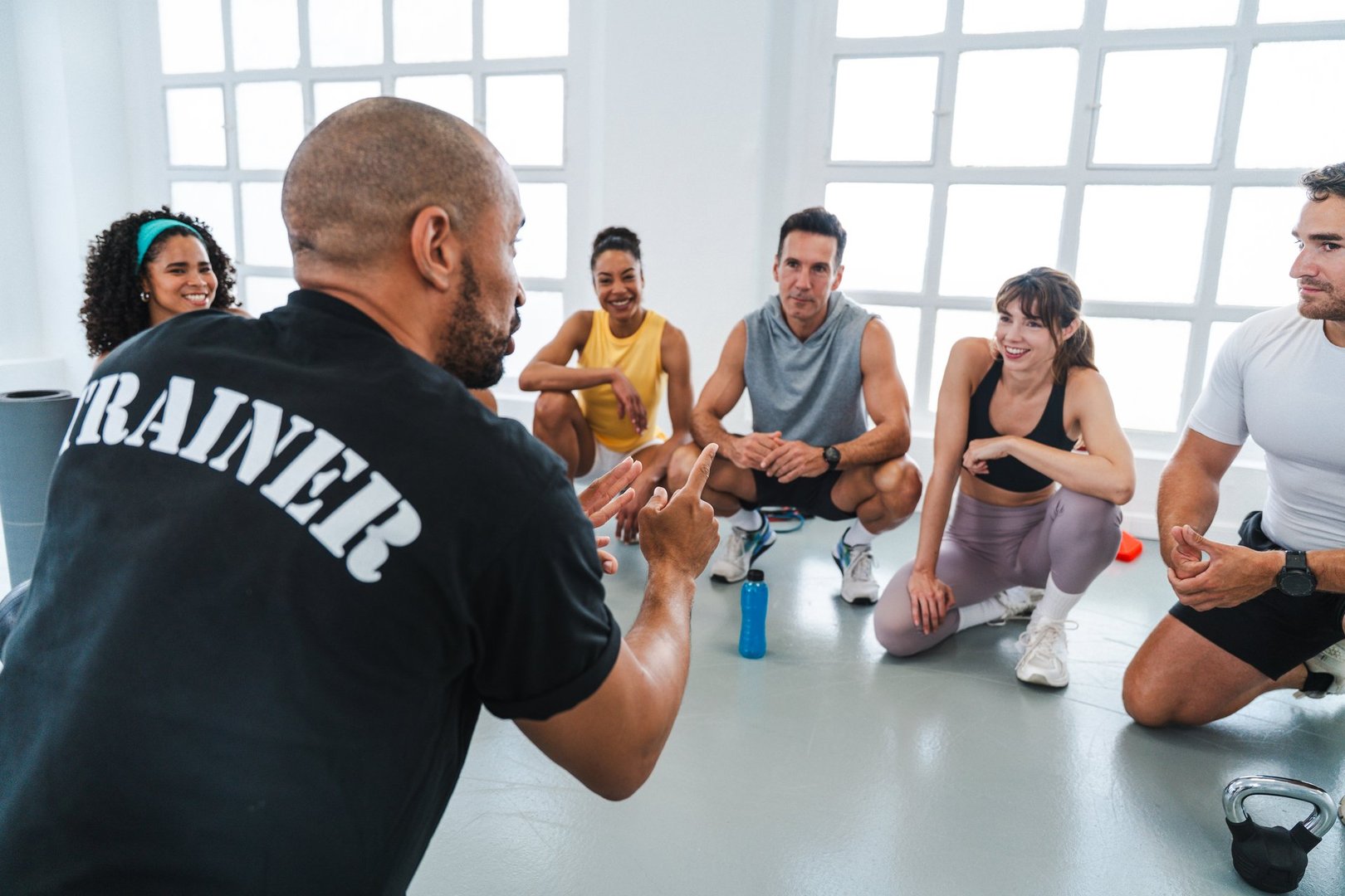 Multiethnic group of sportive people doing functional workout in training class gym personal trainer instructing a group of attentive athletes sitting on the gym floor after a functional workout sessi