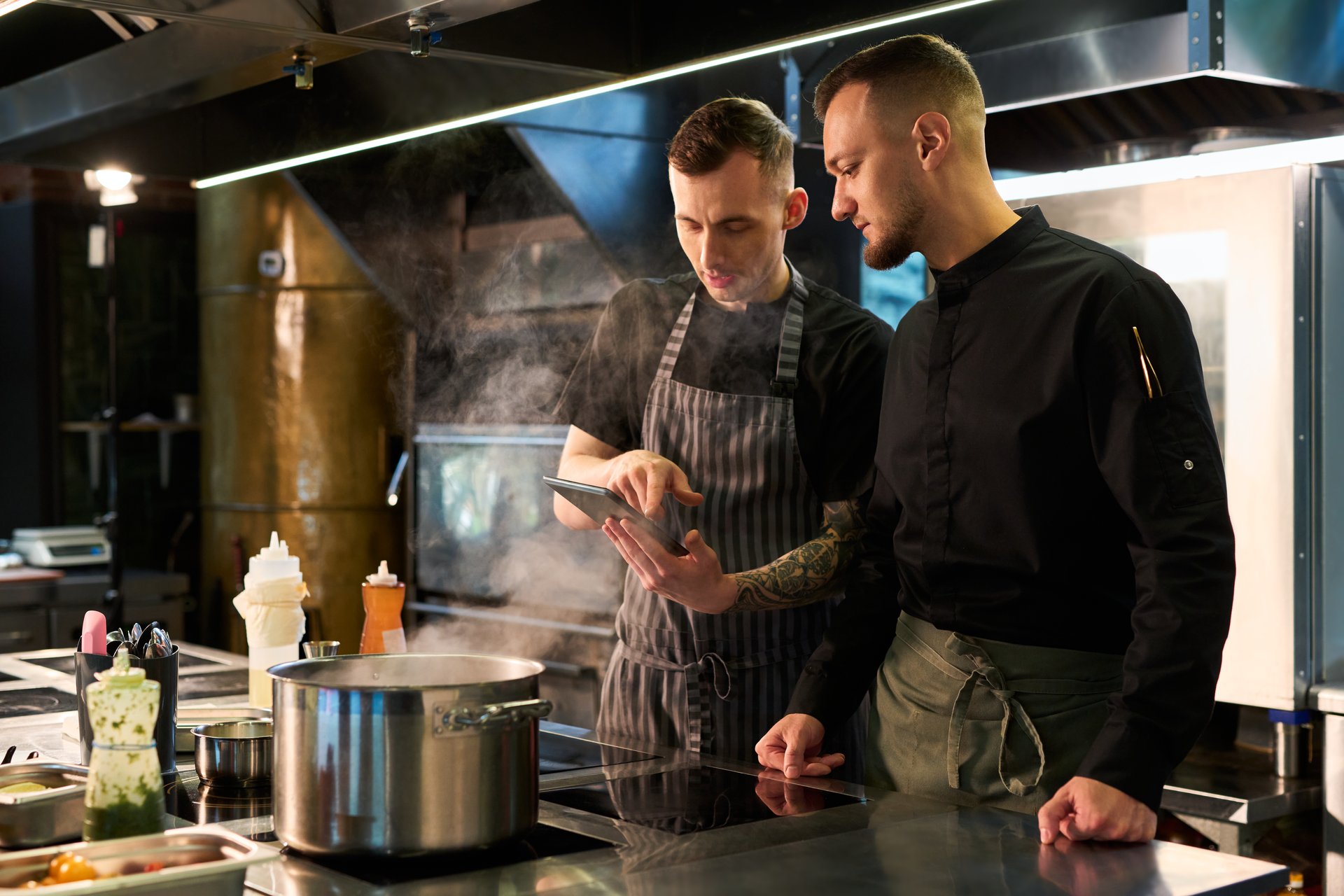 Two young adult Caucasian men standing in professional kitchen discussing recipe while looking at digital tablet near steaming pot, one man wearing apron, both focused on cooking process