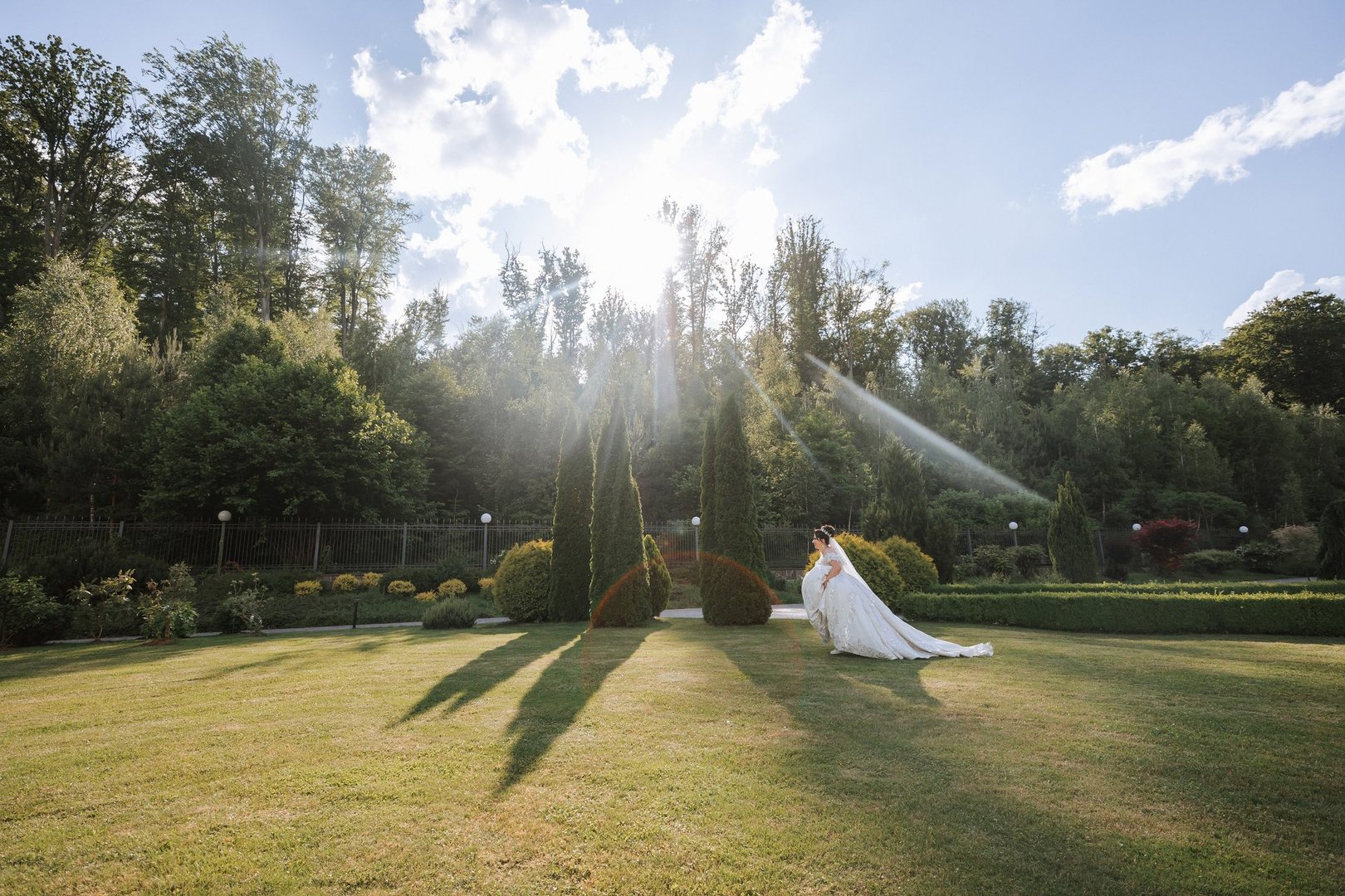 A bride is standing in a field with trees in the background. The sun is shining brightly, casting shadows on the grass. The scene is serene and peaceful, with the bride looking out over the field