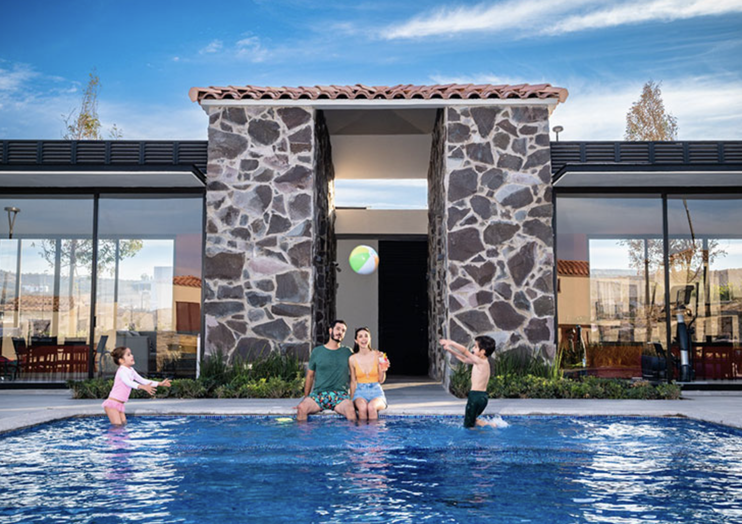 Family playing with a beach ball at the edge of a pool in front of a modern stone building.