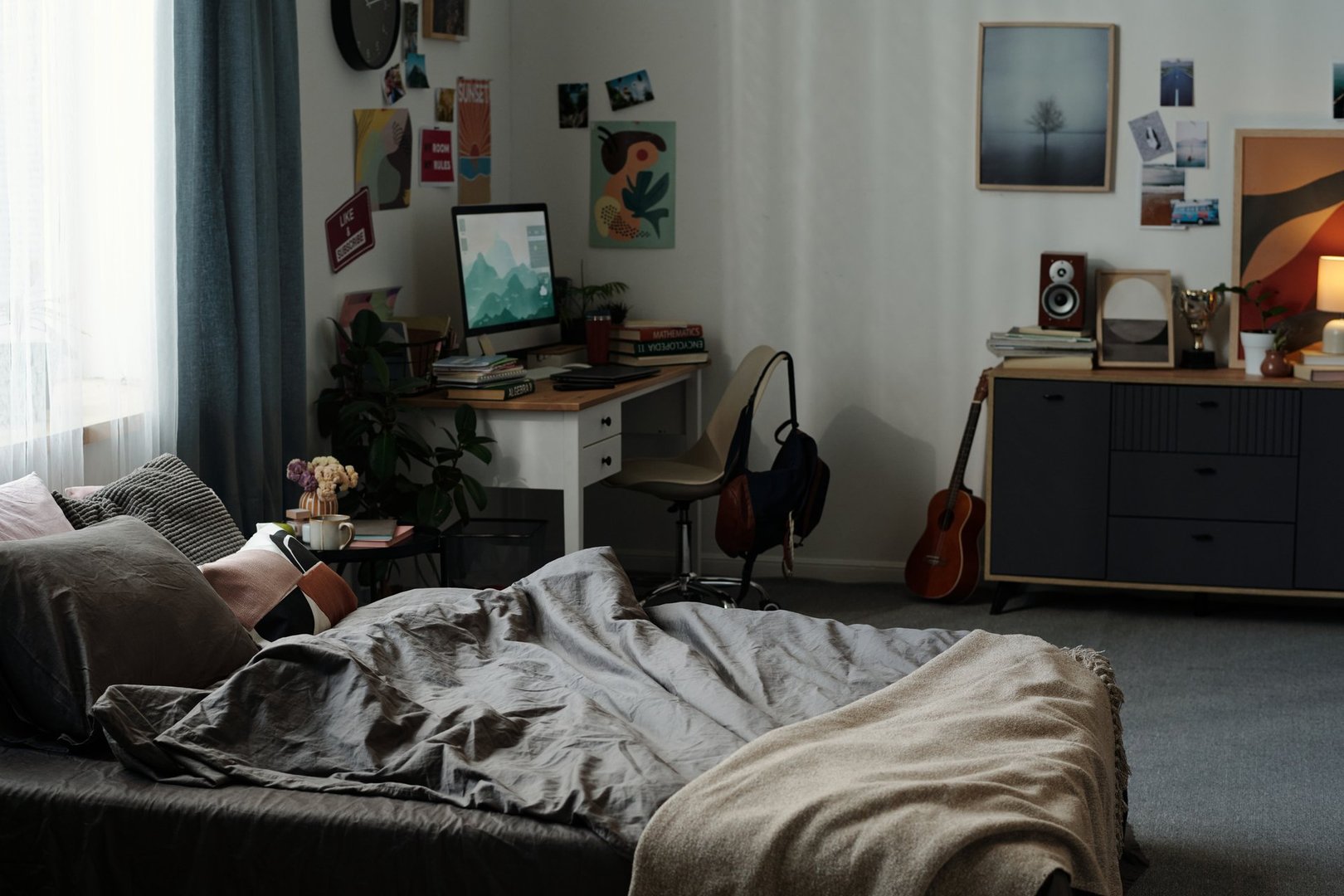 Empty bedroom showing unmade bed in foreground, desk with computer and chair in background, digital devices and cluttered surfaces illustrating concept of gadget addiction