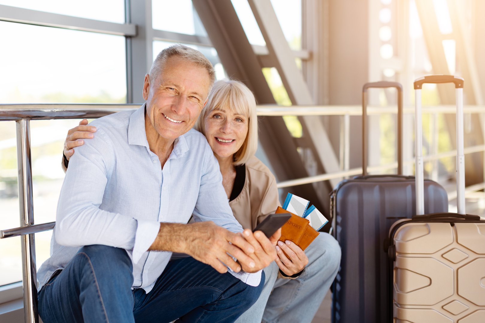 Mature couple sitting at airport with luggage and passports, smiling at camera. Senior family lifestyle travelers enjoying togetherness, love, and holiday freedom