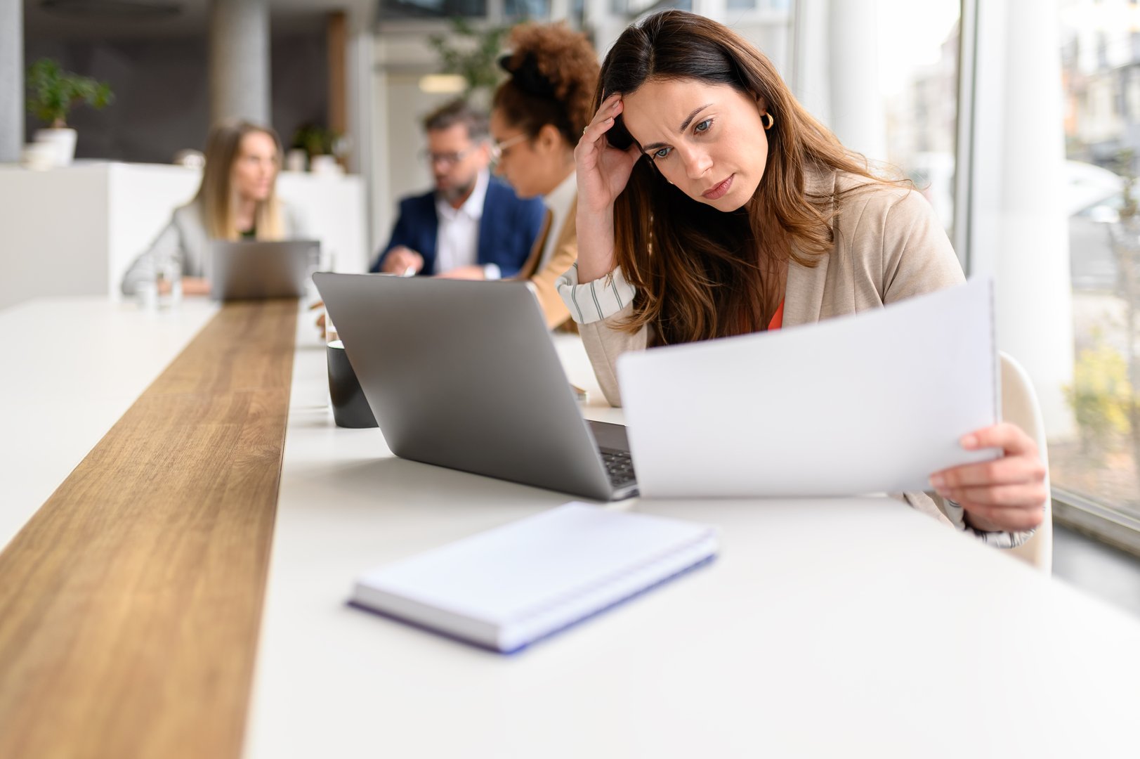 Stressed businesswoman reviewing financial reports while working over laptop in meeting with colleagues in the background