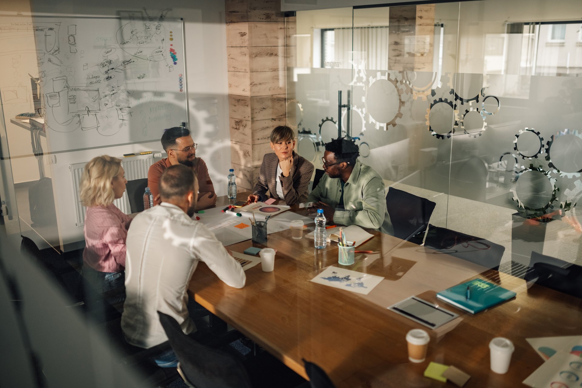 Diverse team of business professionals is having a productive meeting in a modern office, sitting around a wooden table, analyzing charts, and engaging in collaborative discussion