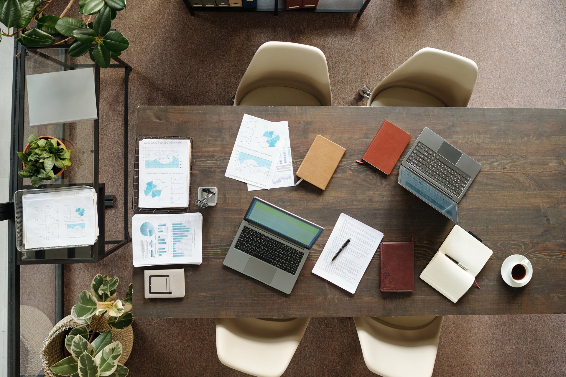 Top view showing workspace with two open laptops, printed charts, notebooks, pen, and potted plants on wooden table, suggesting business meeting or collaborative office work