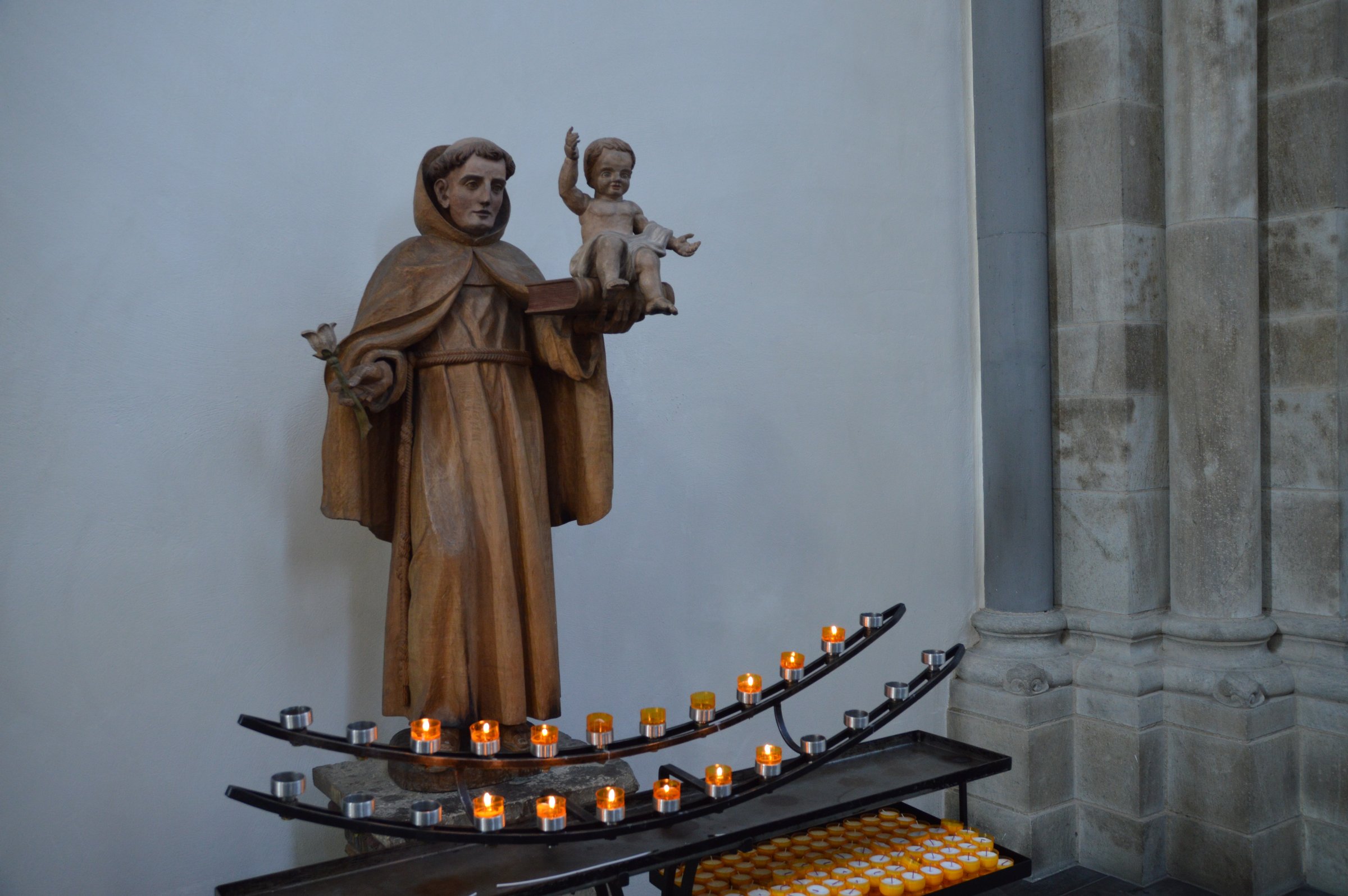 Wooden statue of a saint holding a child, surrounded by lit candles on a stand, inside a church with stone walls.