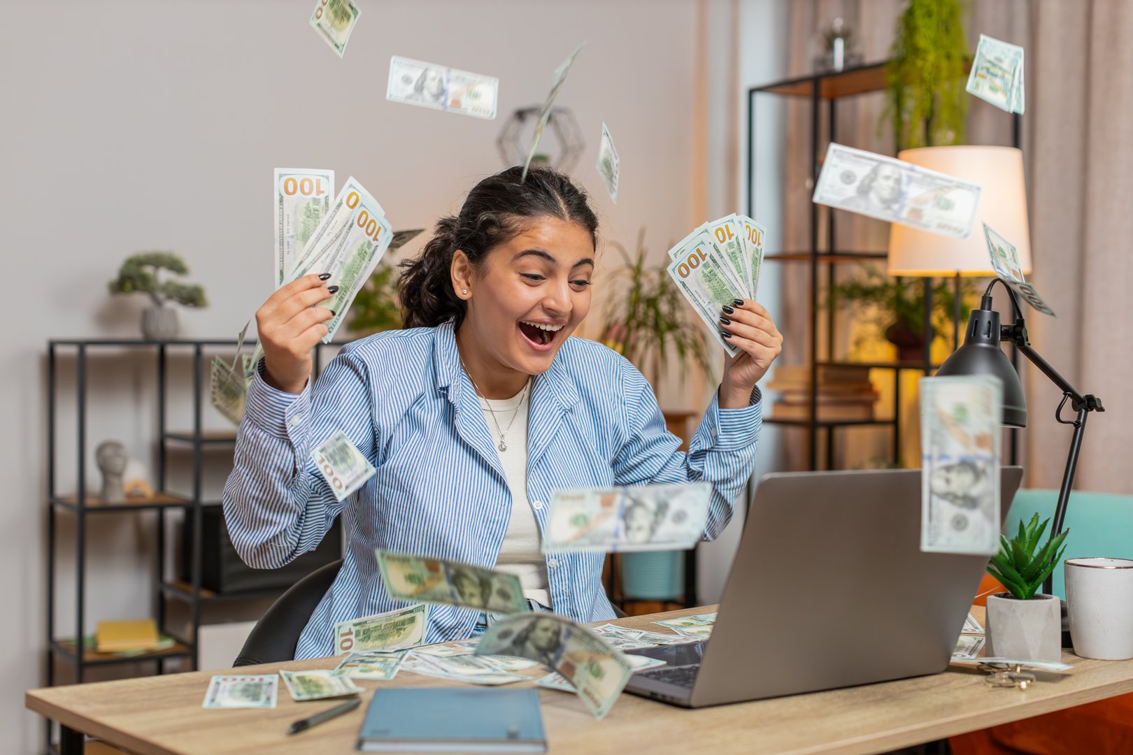 Rich Indian woman earnings big profit success win lottery money cash rain falling shopping more tips. Happy young freelancer girl manager with laptop clenching fists at home office desk. Lifestyles