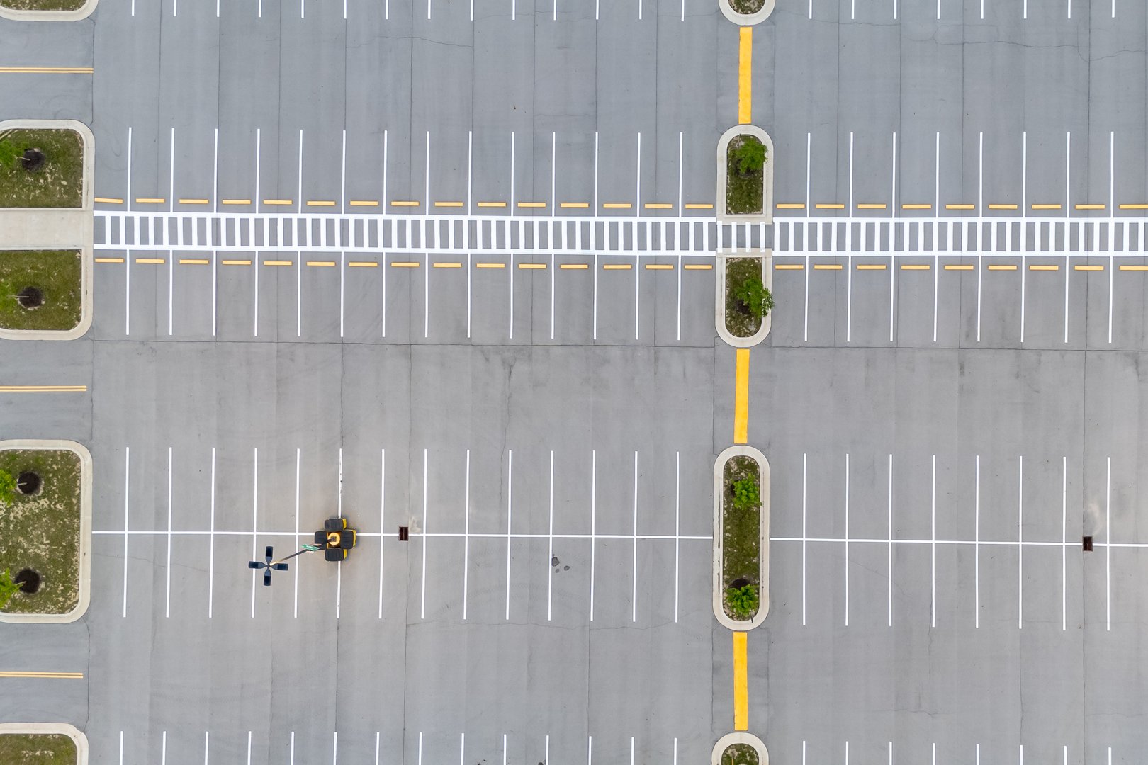 An aerial view reveals a vast, empty parking lot with neatly marked spaces and a central yellow dividing line flanked by small green islands, suggesting a quiet, off-peak moment or a newly completed development.