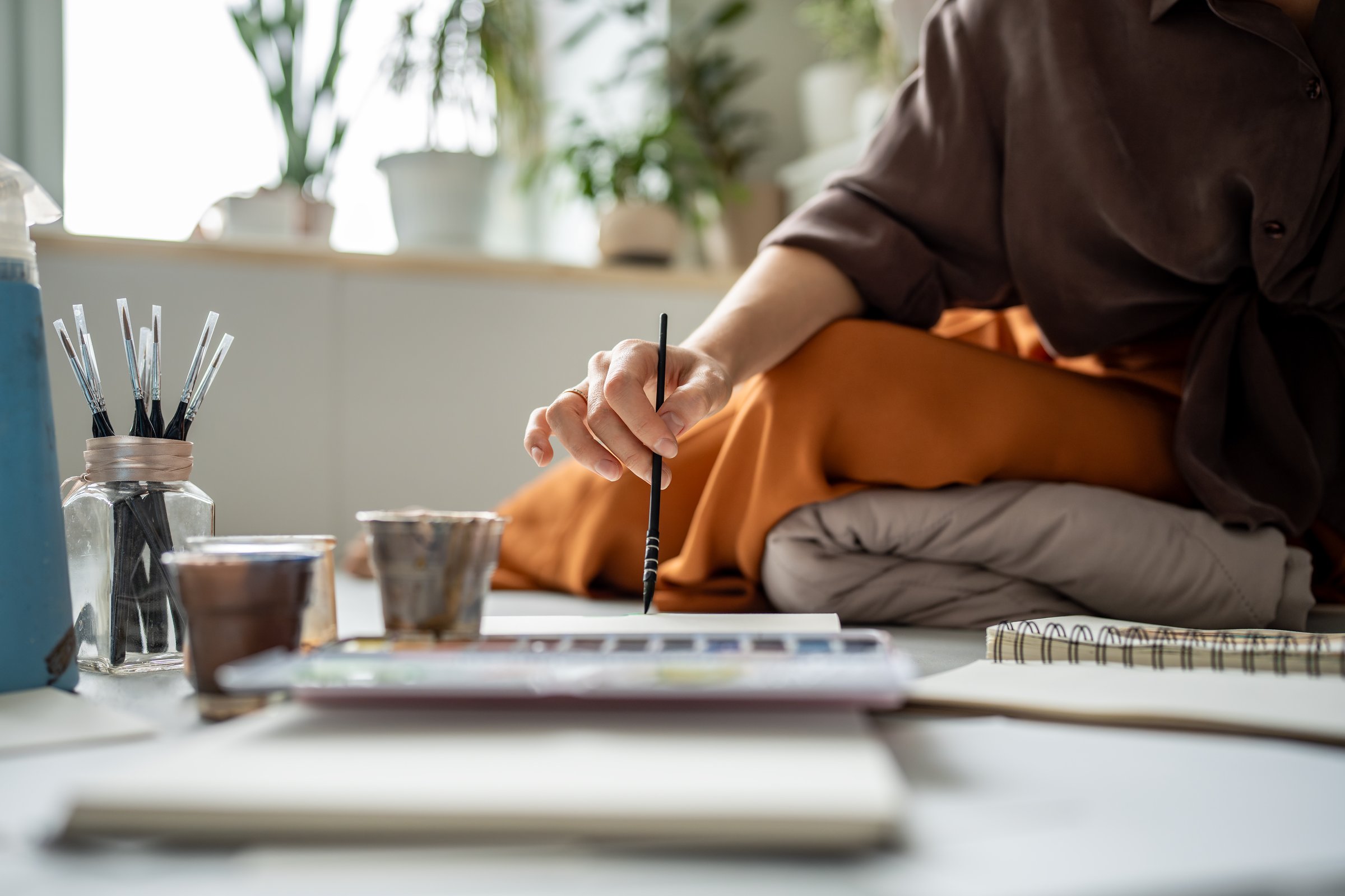 Closeup hand of artist female painting with watercolour in sketchbook holding paintbrush sitting on floor in art studio. Paint tools for creative ideas painters. Hobby for stress relieve, art therapy.