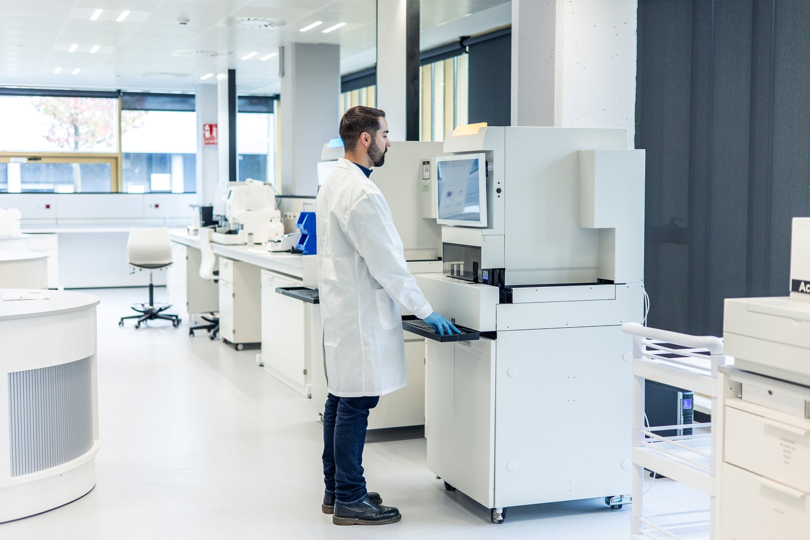 Scientist wearing a lab coat is working with a modern automatic analyzer in a bright and spacious laboratory, performing tests and analyzing samples