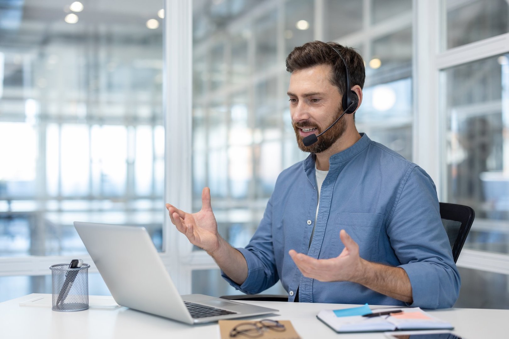 Young man with headset talking to colleagues and partners remotely using laptop for video call. Support worker, helpline consultant inside office.
