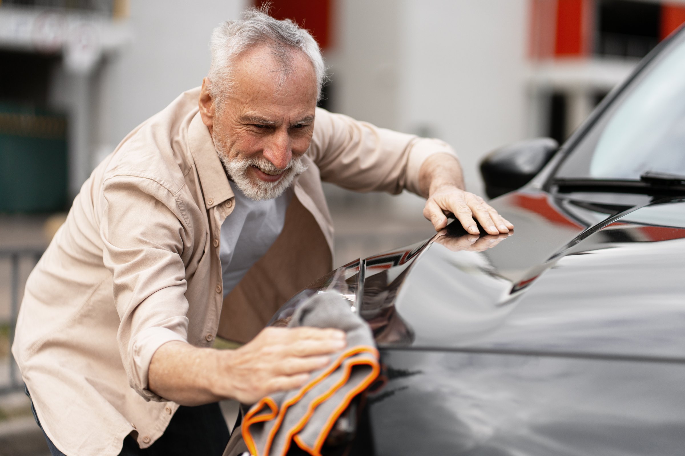 Happy senior man cleaning his car with microfiber cloth outdoors, enjoying his retirement and taking care of his vehicle with care and attention. Driving concept