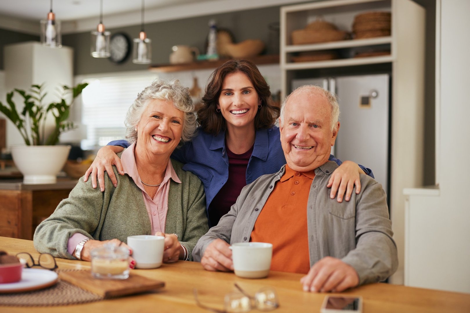 Portrait of old satisfied father and joyful proud mother sharing tea and heartfelt conversation with adult daughter during tea time. Beautiful daughter embracing from behind her elderly parents while smiling together and looking at camera. Warm family moment with senior mother and elderly father enjoying tea together.