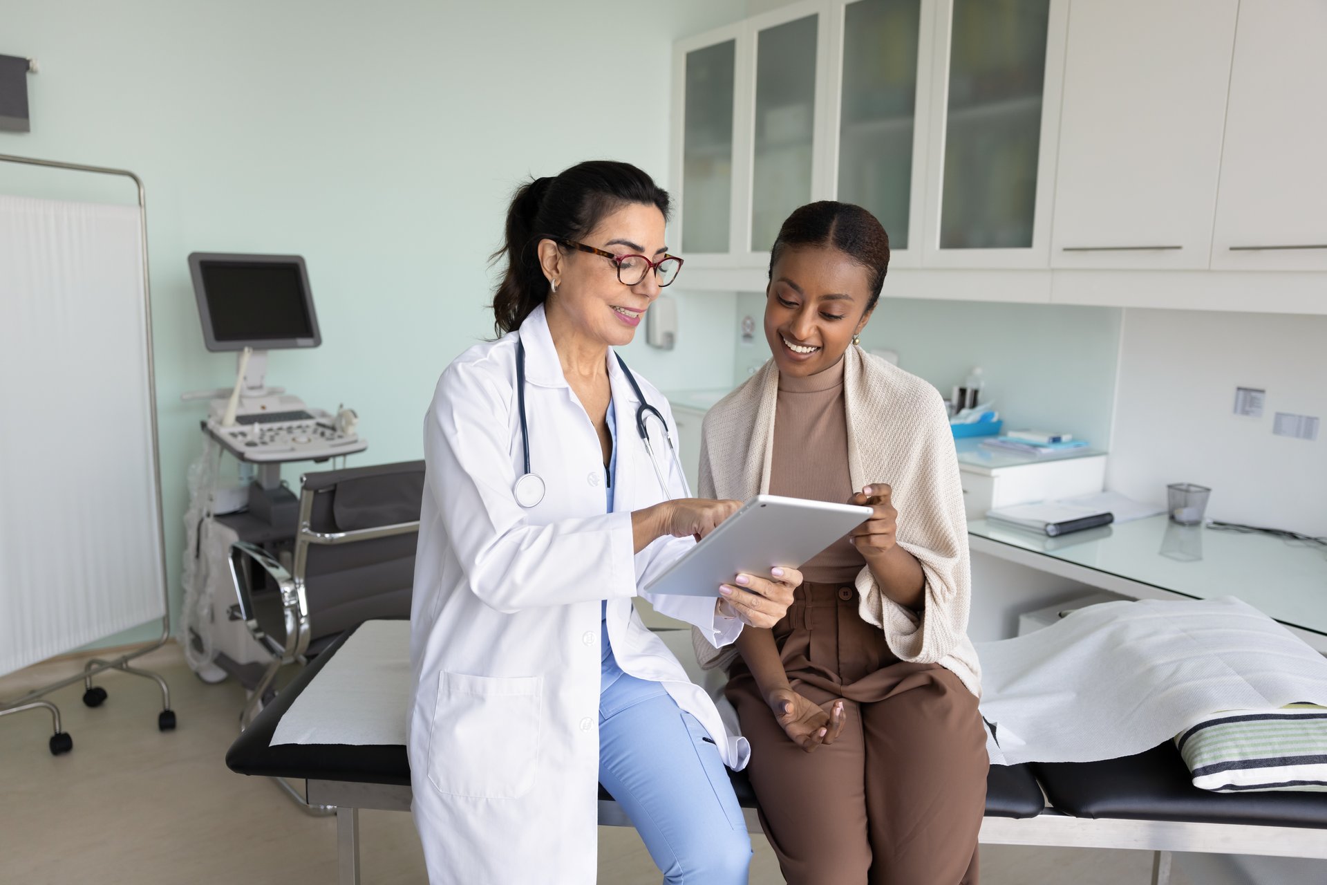 Positive senior ultrasonography doctor showing ultrasound examination results to young African female patient, explaining medical checkup electronic result, telling good news, smiling