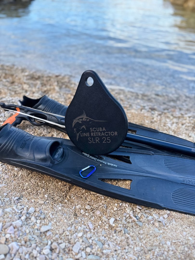 Pair of black scuba fins with a Scuba Line Retractor SLR 25 device on a pebble beach near water.