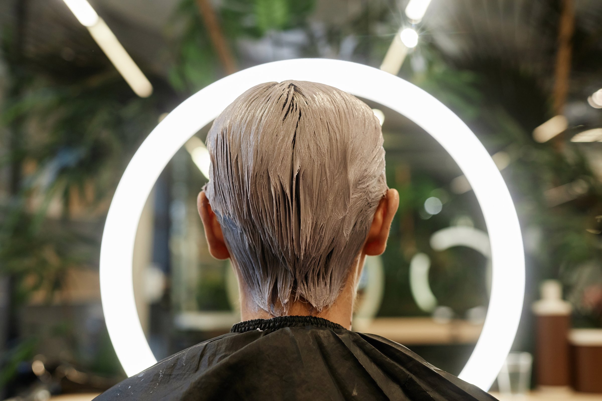 Back view of young man with bleached hair facing ring light in beauty salon and taking photos