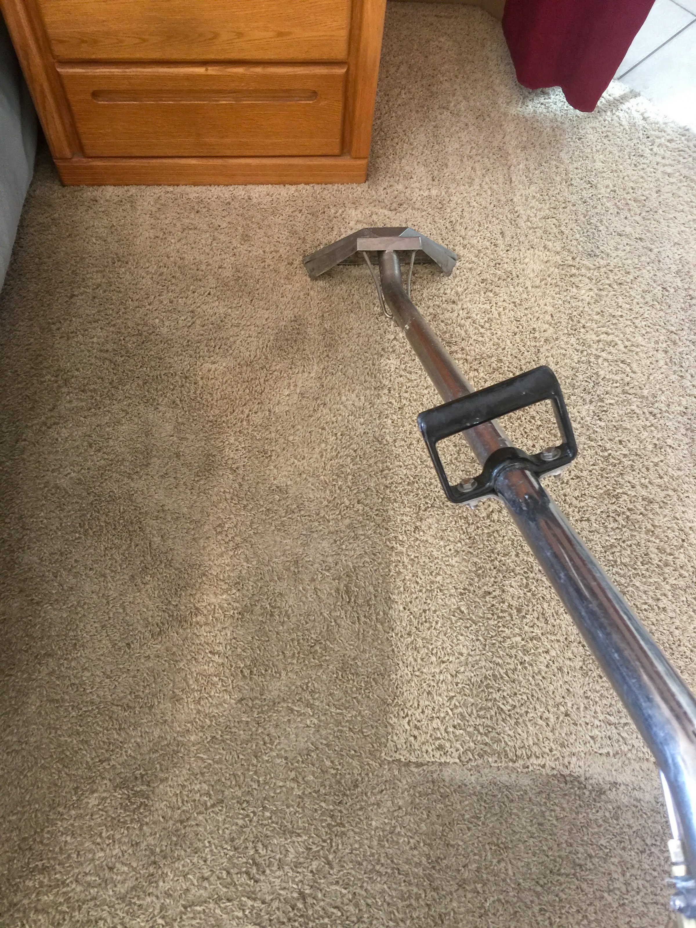A detailed image of a professional carpet cleaning tool in use, showing a dramatic difference between a cleaned and uncleaned area of beige carpet. The process is taking place near a wooden dresser in a home interior. This image emphasizes the efficiency of steam or deep carpet cleaning services, ideal for promoting cleaning businesses, home improvement, or cleanliness concepts.