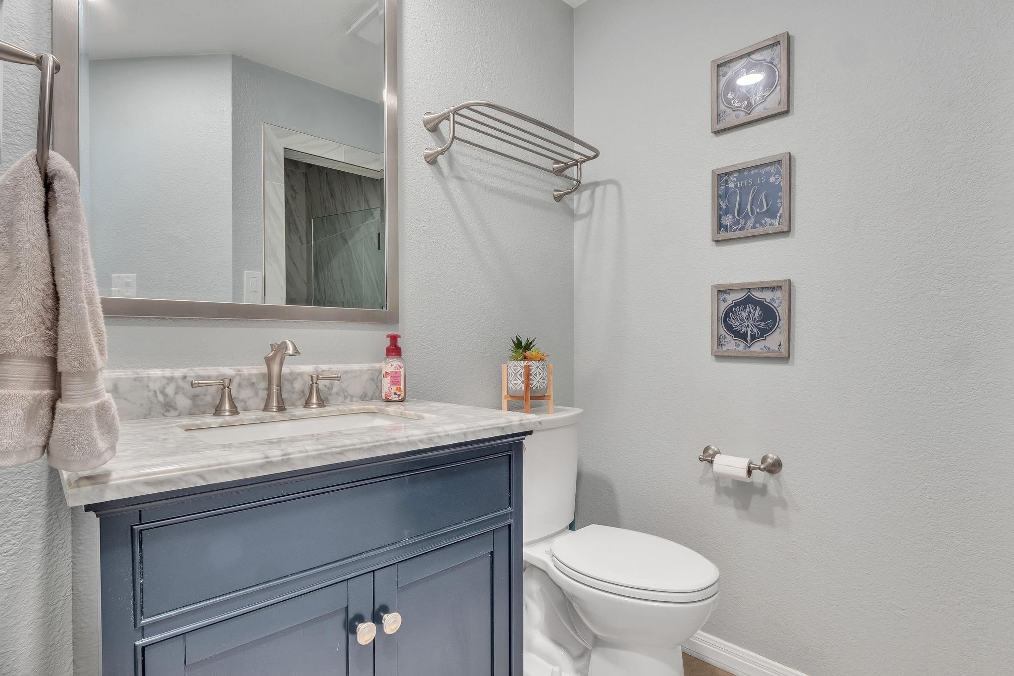 Bathroom with blue vanity, marble countertop, towel rack, toilet, and framed art on light gray walls.