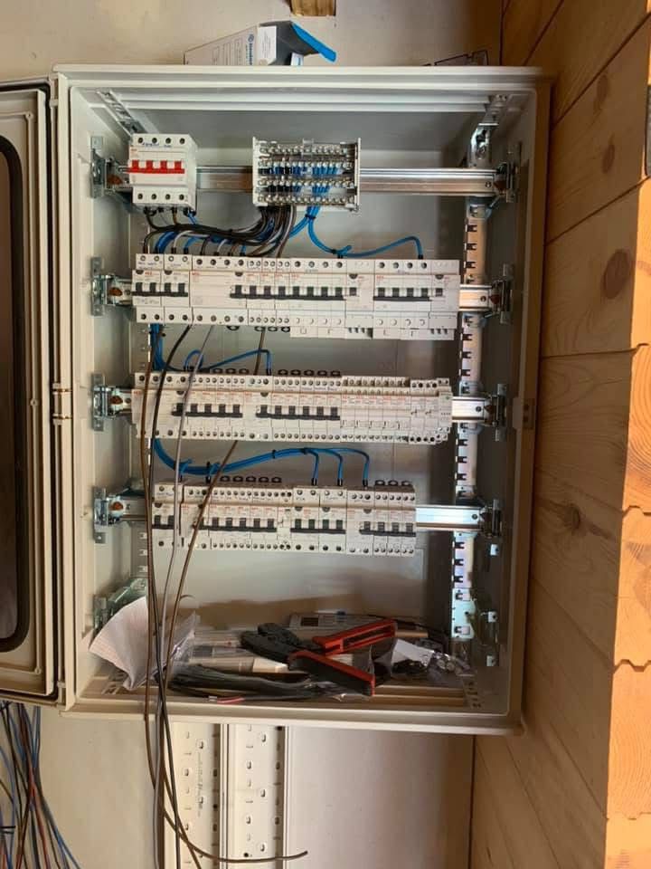 Electrical panel with circuit breakers and wiring, tools inside, mounted on a wall with wooden boards visible.