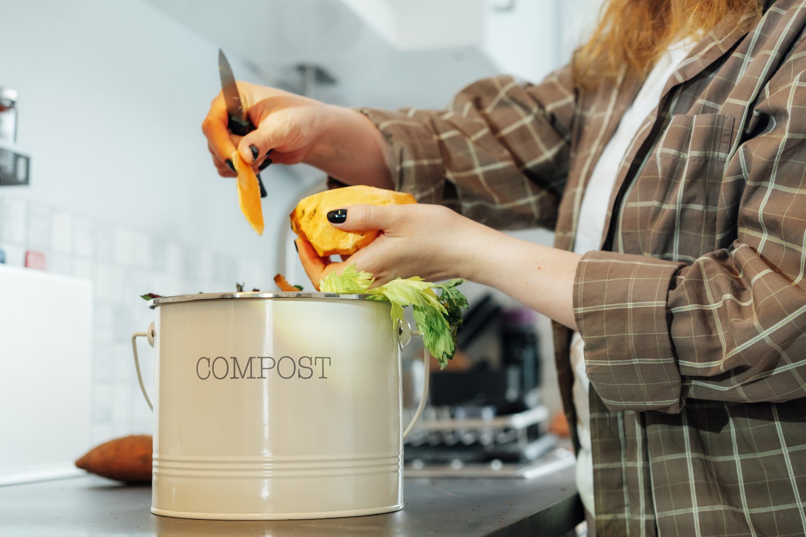 Compost the kitchen waste, recycling at home. Compost bin with vegetables cutted leftovers on the kitchen table with blurred woman cooking meal. Environmentally responsible, ecology. Selective focus.