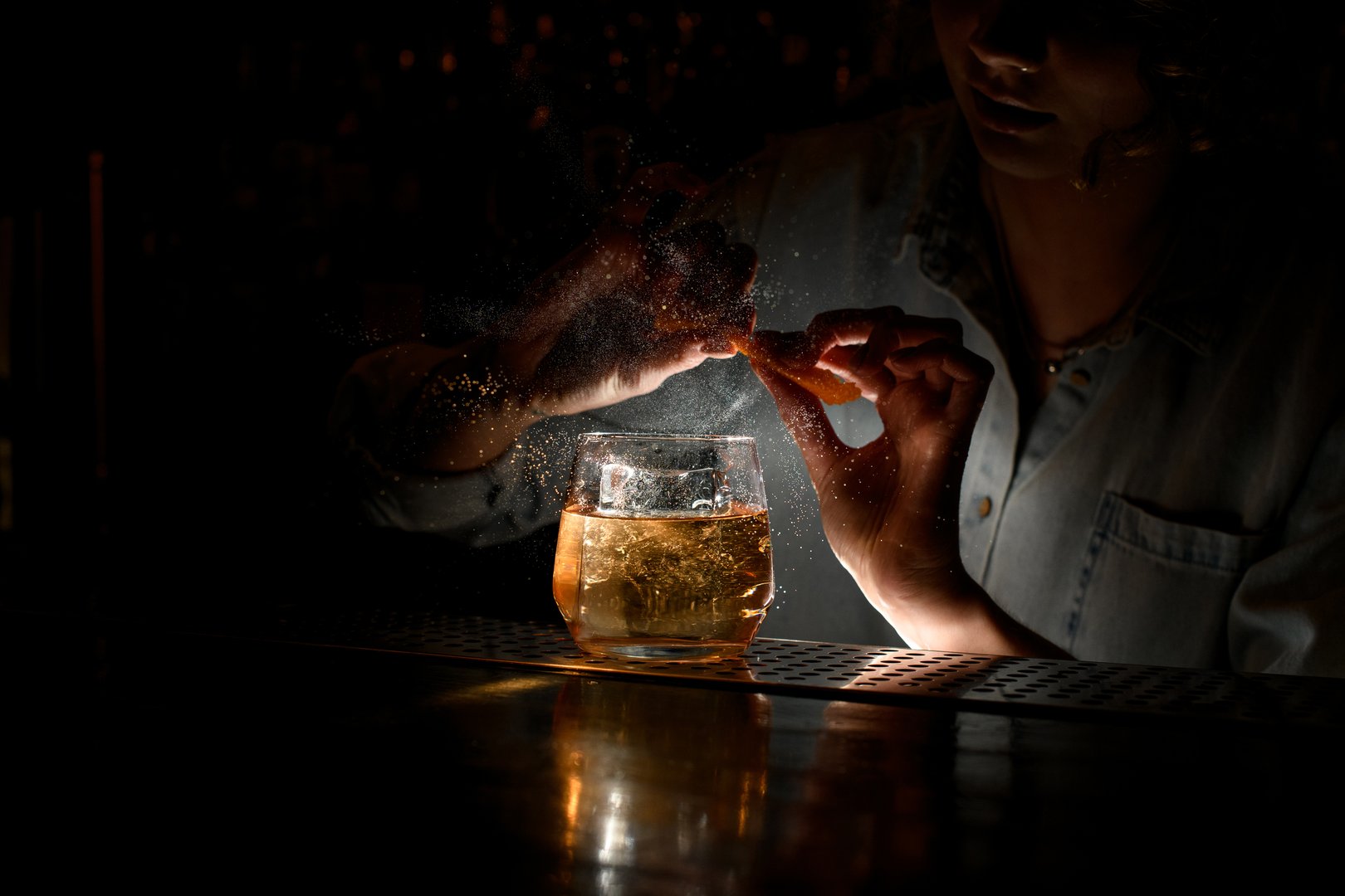 woman bartender at dark bar neatly sprinkles citrus juice on glass with cocktail on bar counter.