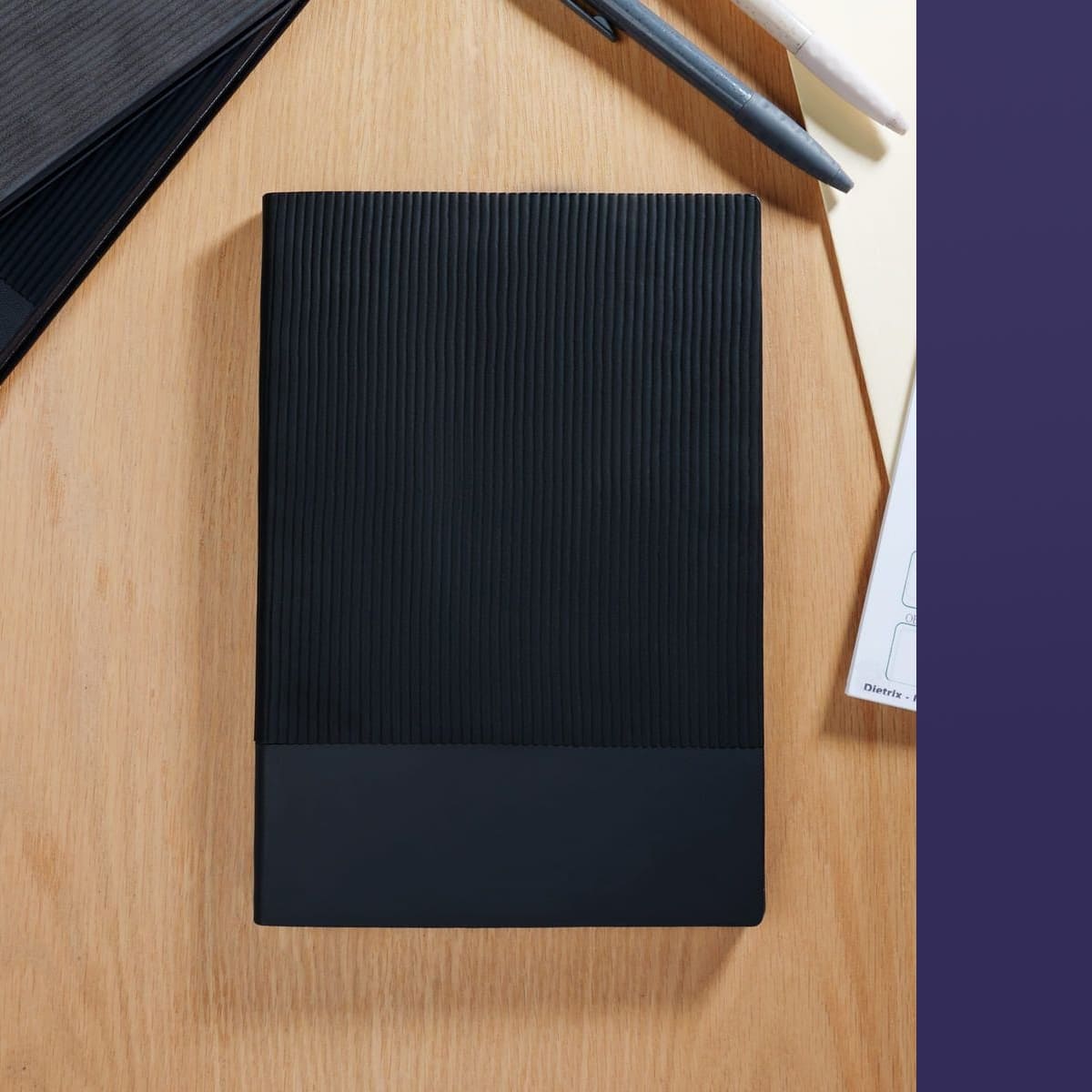 Black notebook on a wooden surface with a purple side panel and two pens nearby.