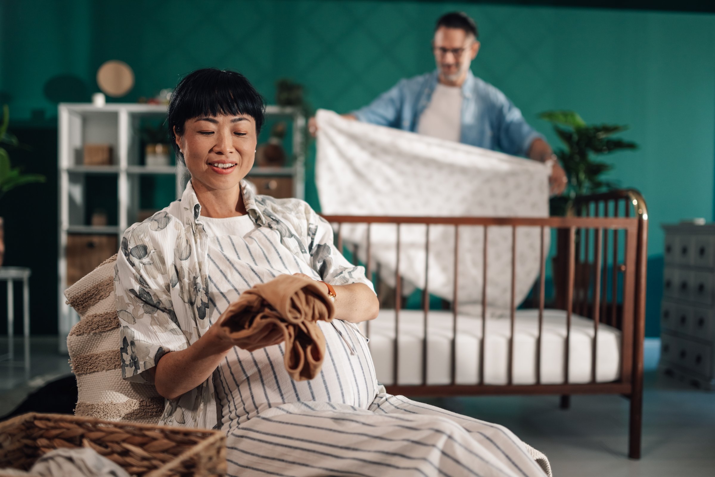 Happy pregnant asian woman folding baby clothes, while blurred man is preparing crib in background