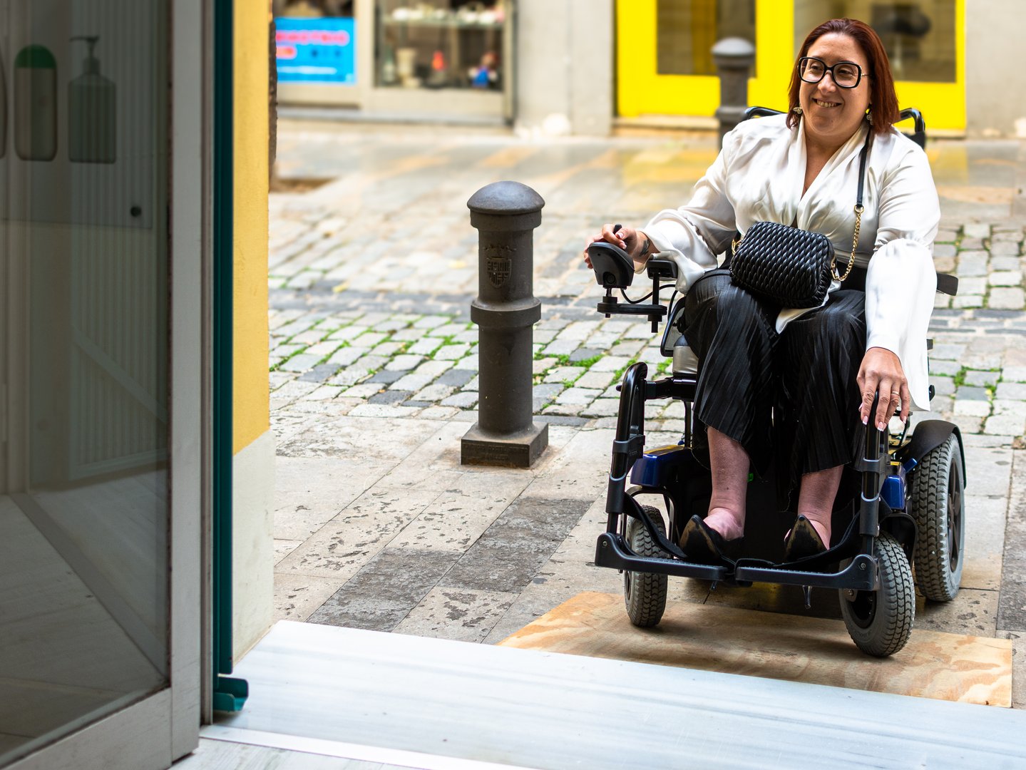 A woman in an electric wheelchair smiles as she enters a store using a ramp, demonstrating accessibility in retail.