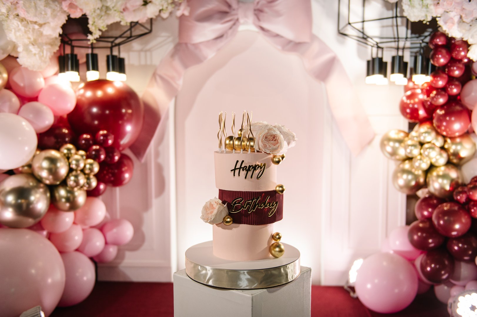 Trendy burgundy Cake with candles on background balloon and big bow on arch. Birthday party decorations. Three-tiered cake with pink roses, Happy Birthday text topper, gold decor. Celebration concept.