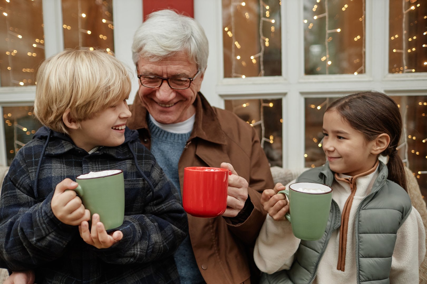 Senior Caucasian man smiling while sitting with boy and girl, holding mugs and interacting together in village setting, children looking at man with joy