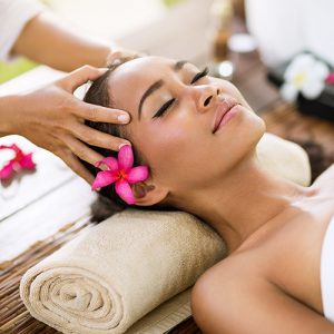 Person receiving a relaxing head massage, lying on a towel with a flower in hair, eyes closed, serene expression.