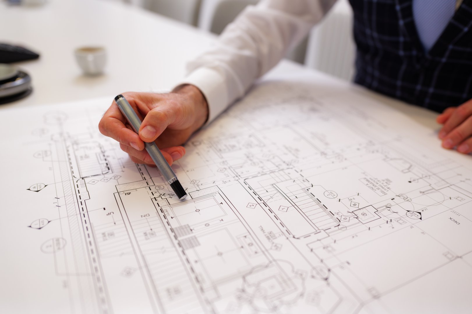 Architect is carefully reviewing a blueprint spread out on a white table, using a pen to point at specific details of the architectural plan