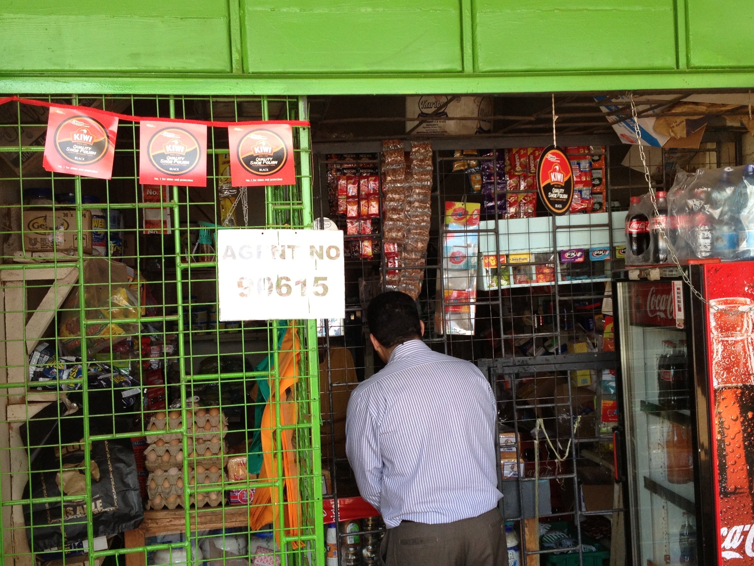 A man stands at a small grocery store counter, surrounded by various products and drinks, with a green awning above.