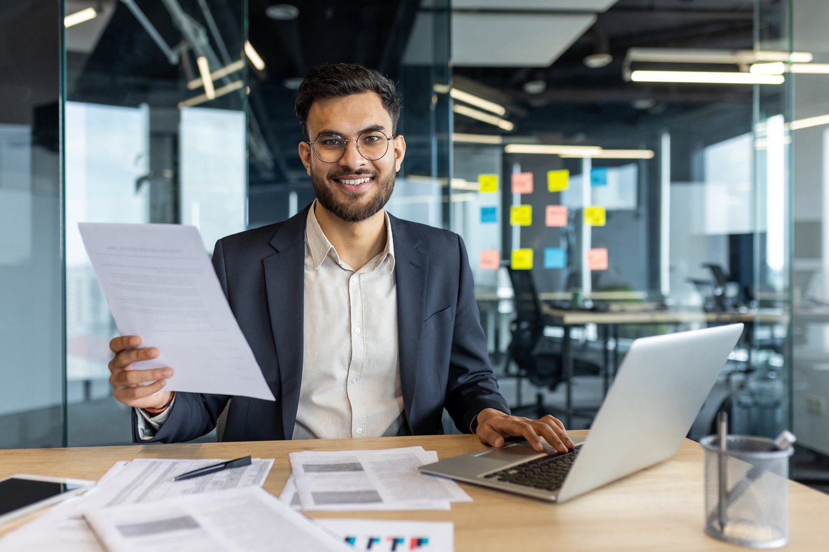 Mature male businessman examining documents and using a laptop at his desk in a modern office. He appears confident and professional while focusing on work.