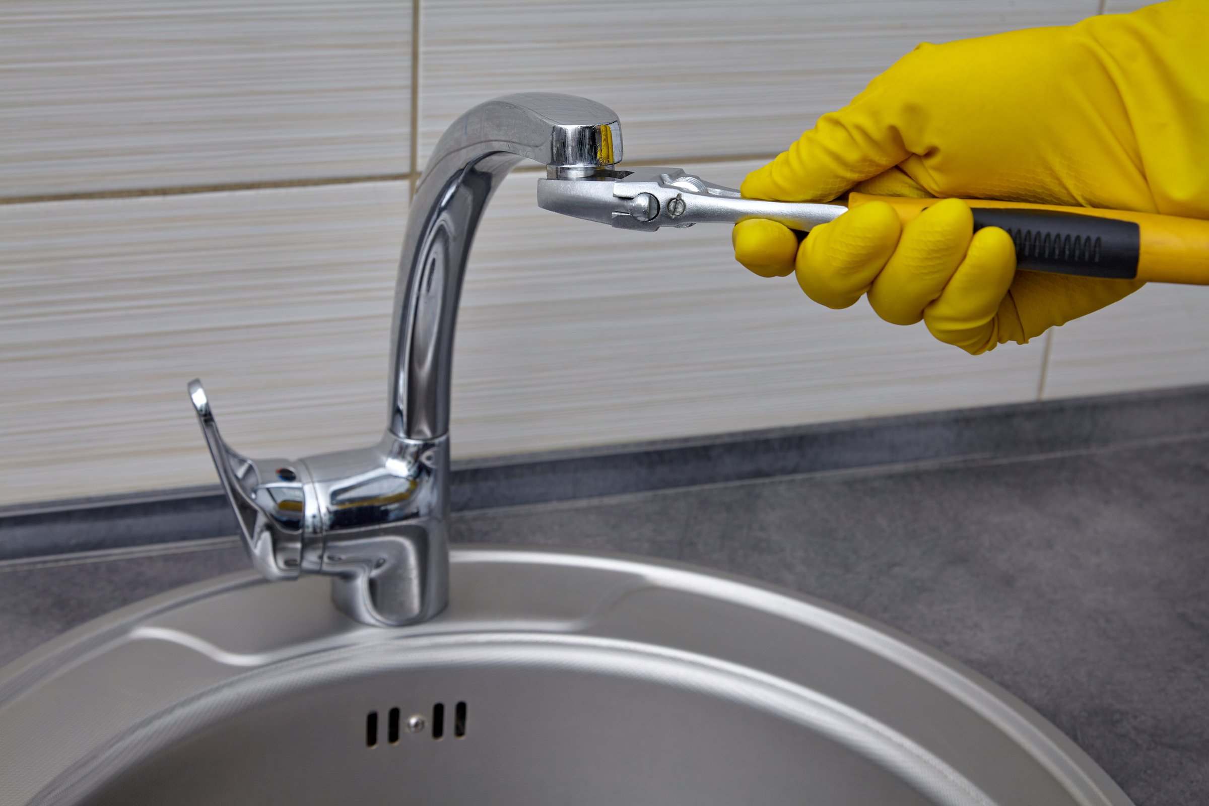 A plumber in yellow rubber gloves unscrews the faucet aerator with an adjustable wrench.