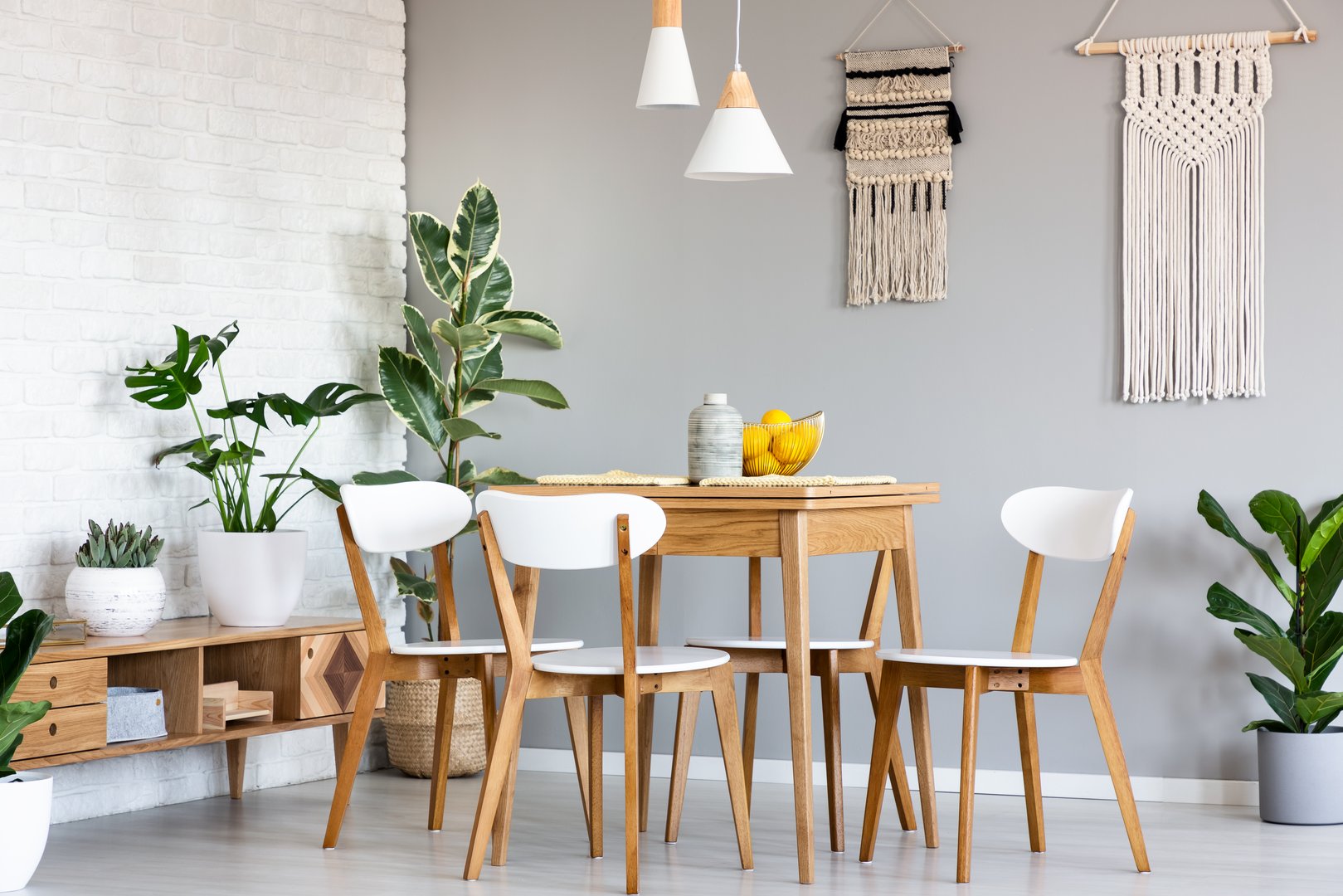 Macrame hanging on gray wall above wooden table and chairs in bright dining room interior with lots of plants. Real photo