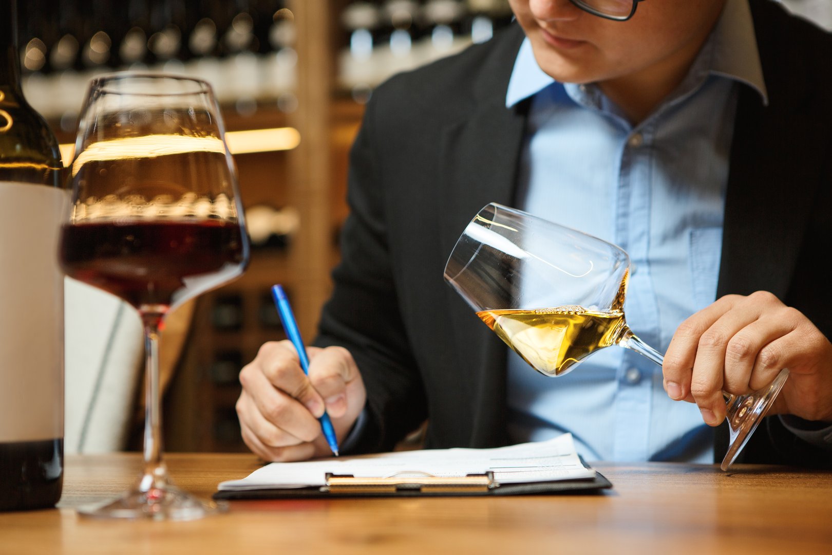 Close-up photo of wine expert tasting white wine in glass and making notes while sitting at table in cellar surrounded by shelves with wine bottles. Professional wine tasting and evaluation concept. Design for poster, banner, postcard.