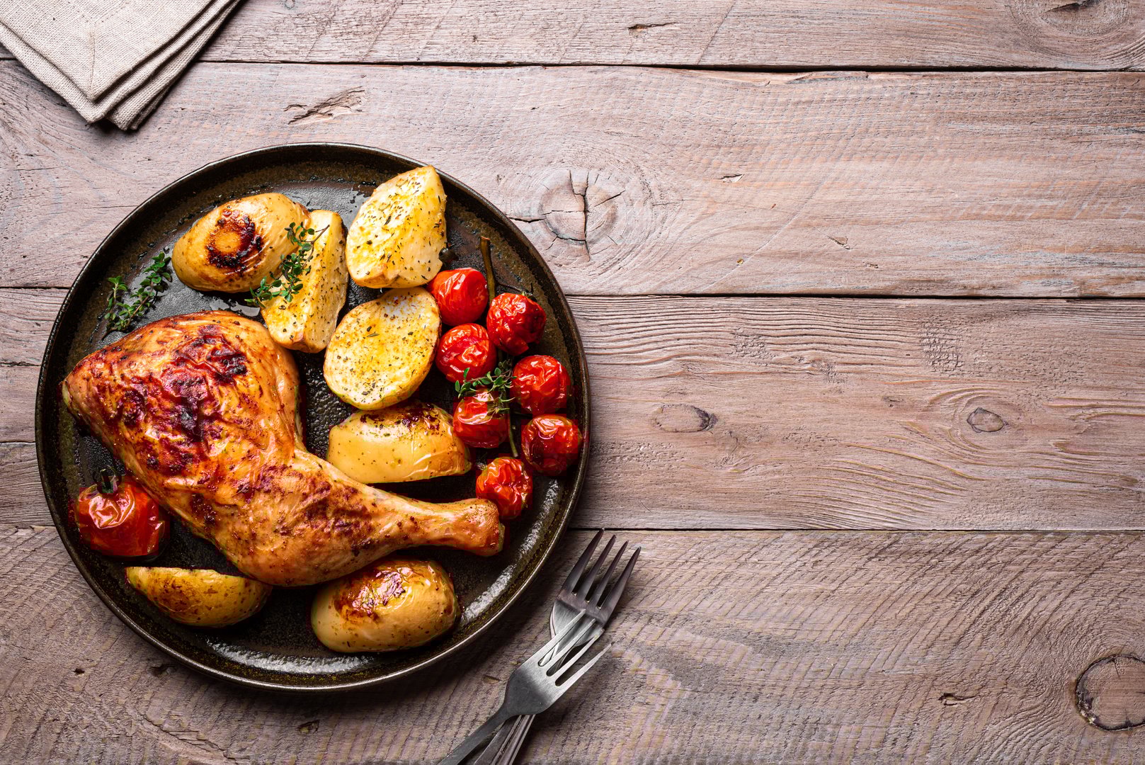 Oven roasted chicken leg quarter with potatoes, tomatoes and herbs served on wooden background, top view, copy space. Hearty homemade meal - chicken and vegetables.