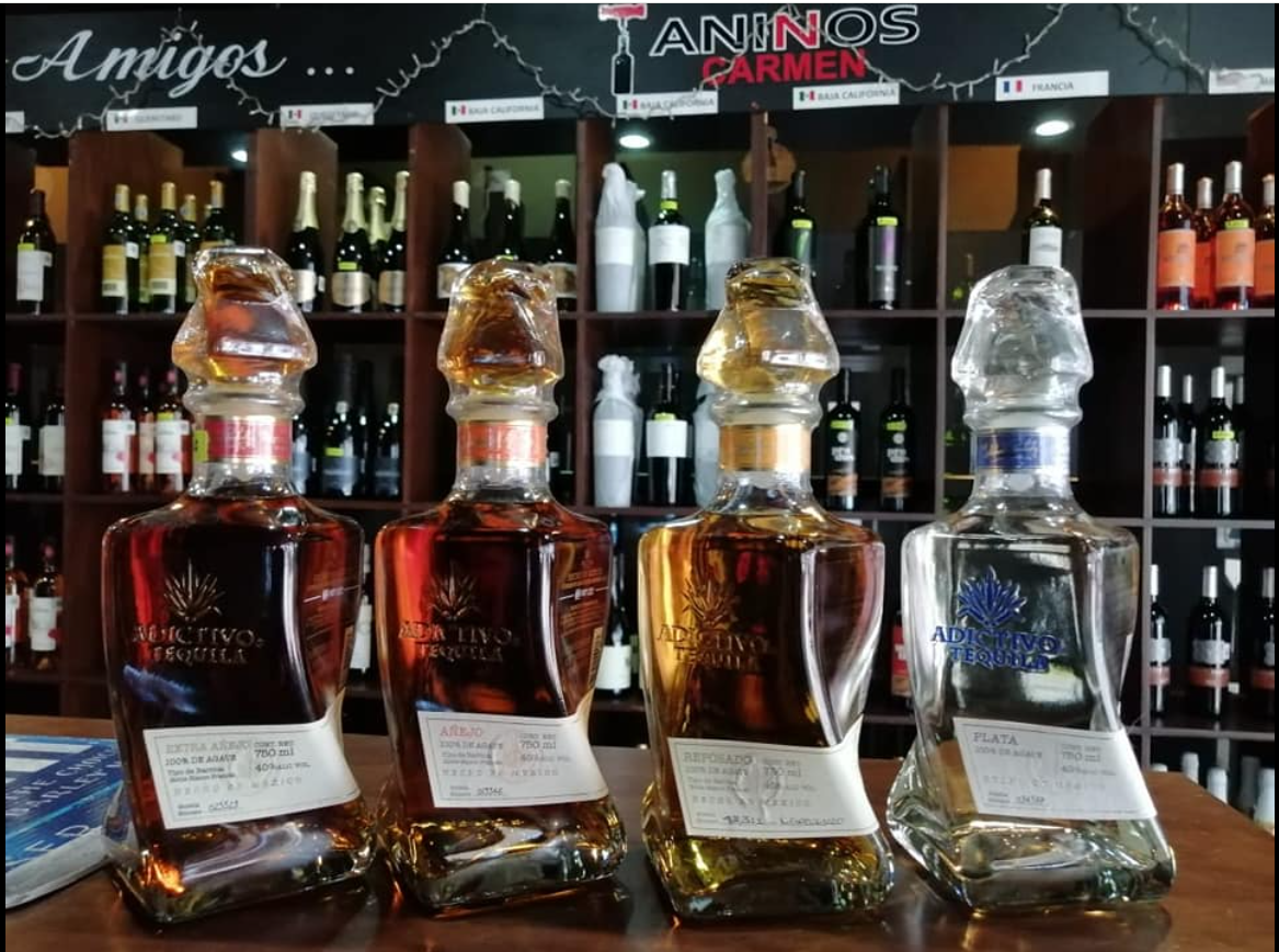 Four bottles of Adictivo Tequila on display in front of a wine shelf with various bottles in a liquor store.