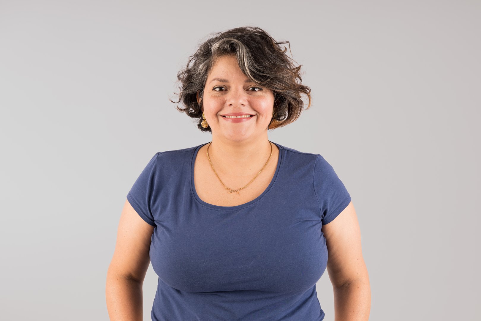 A woman with a blue shirt and a gold necklace is smiling. She has a lot of hair and a big bust