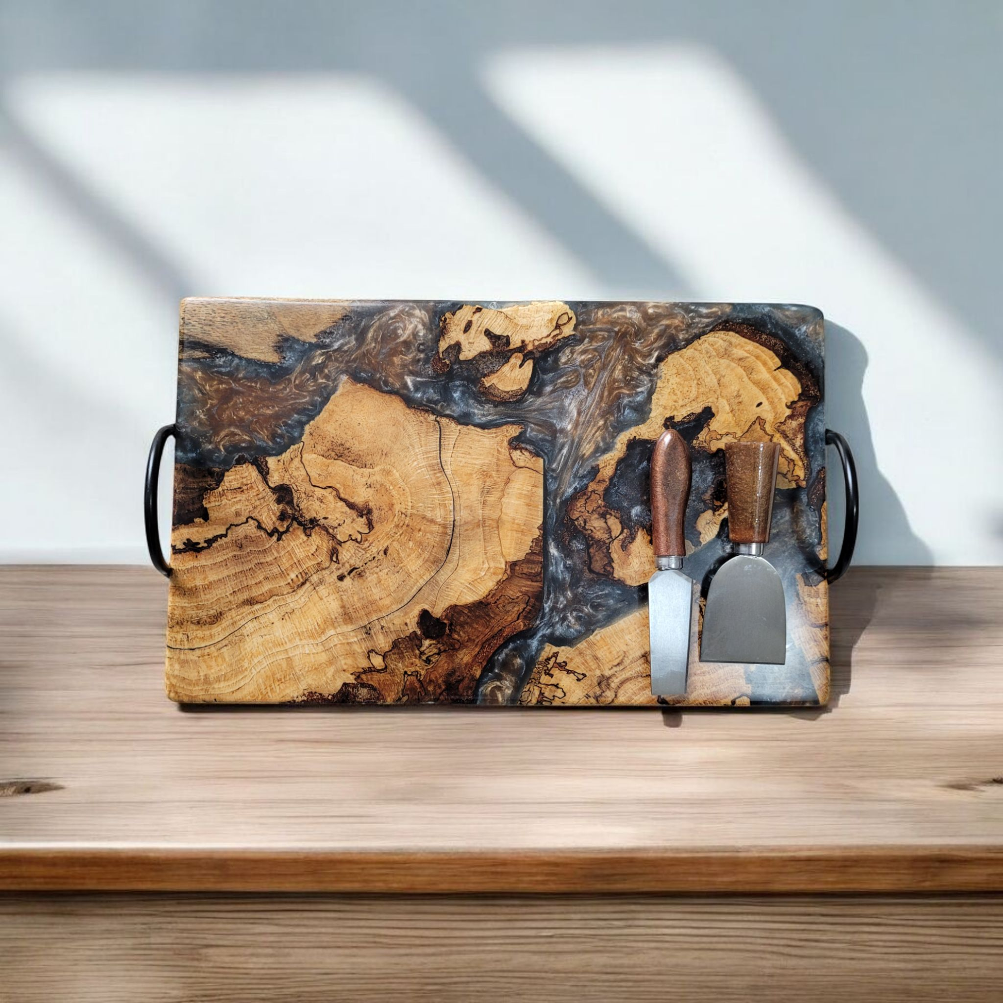 Wooden serving tray with metal handles and two cheese knives, featuring a marbled wood and resin design, on a wooden surface.