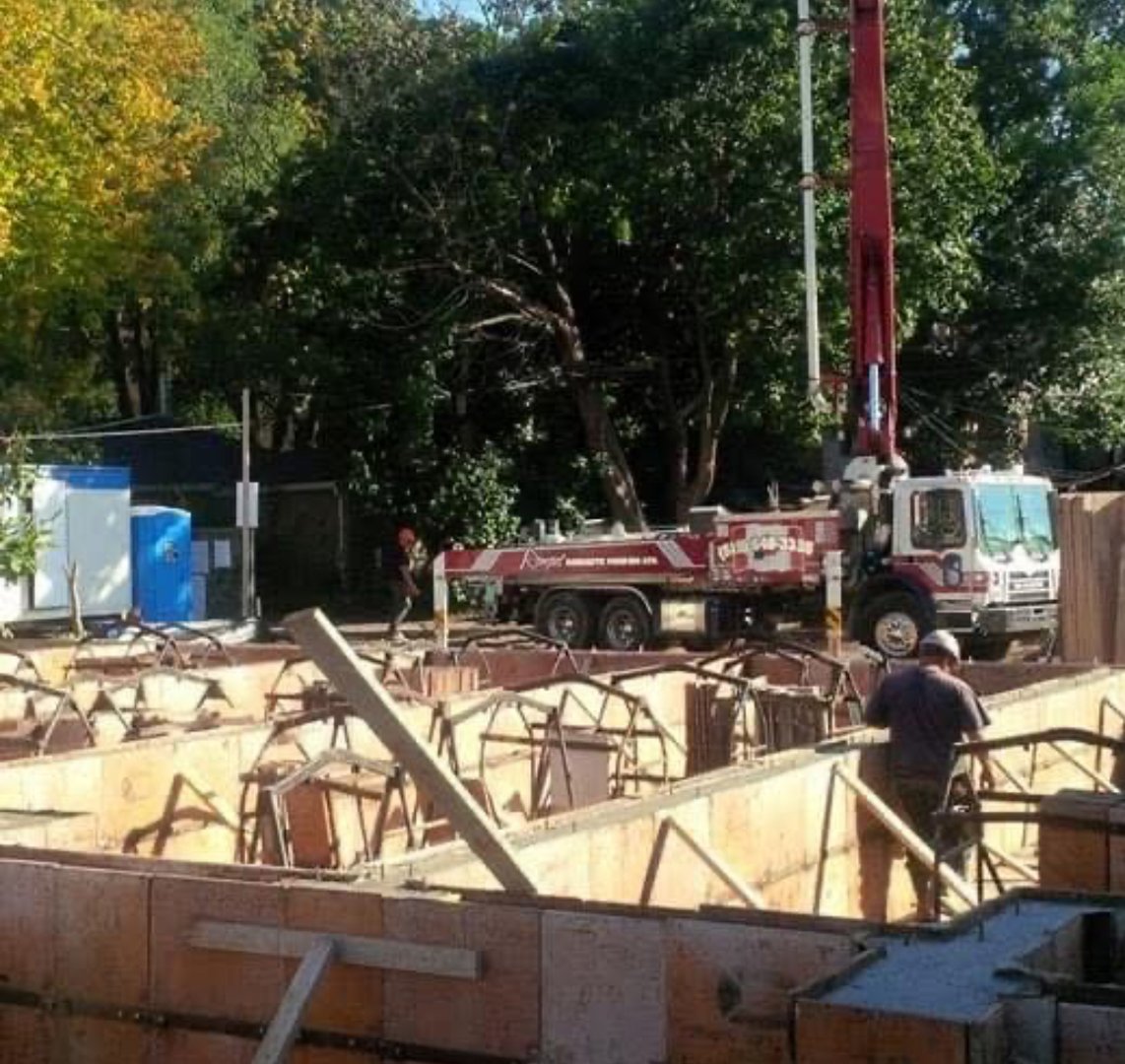 Construction site with wooden frames, person working, and crane truck; trees visible in the background.