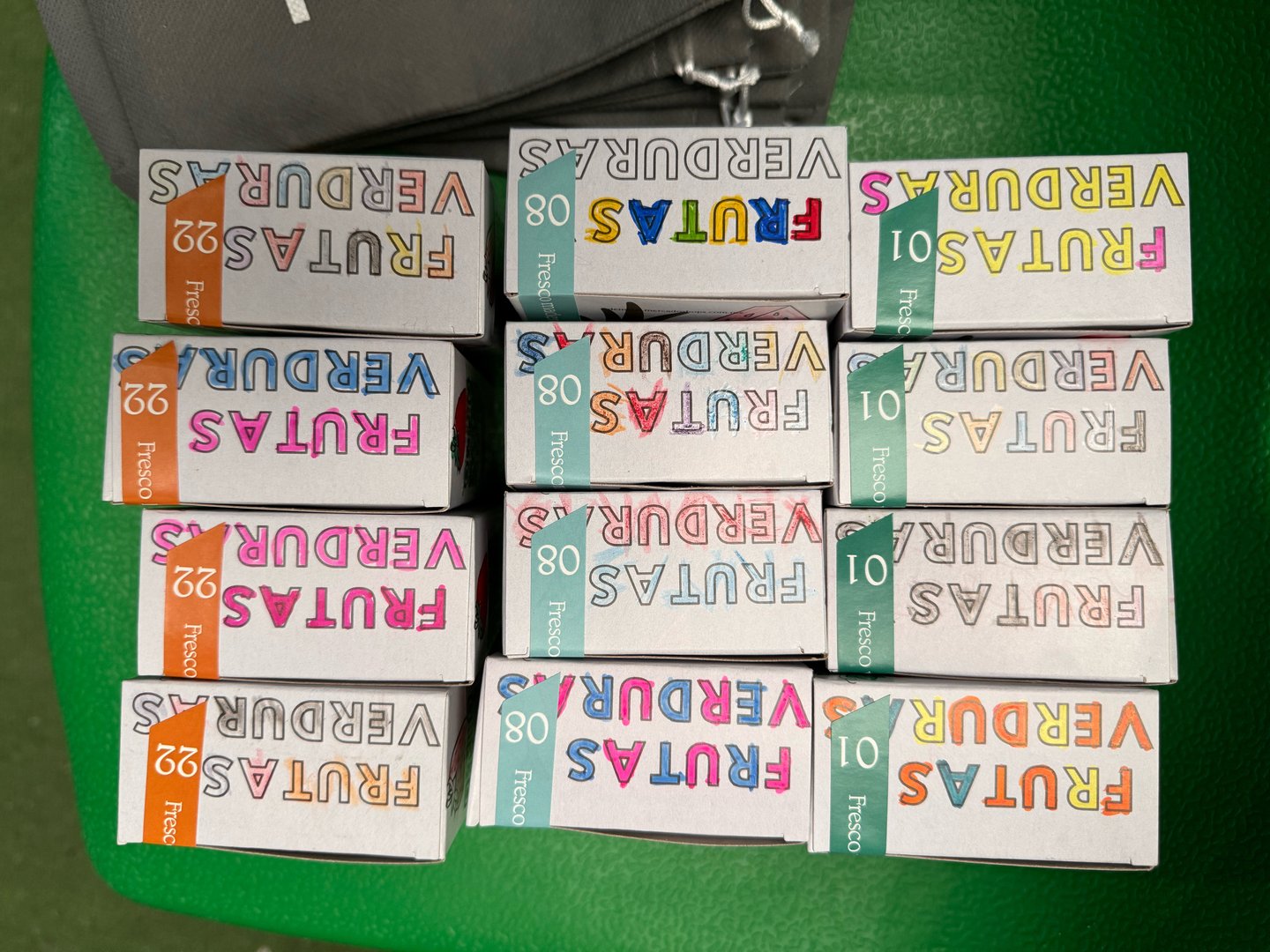 Boxes of colored pencils labeled "FRUTAS VERDURAS" with numbers 36, 80, 50, and 10, arranged on a green background.